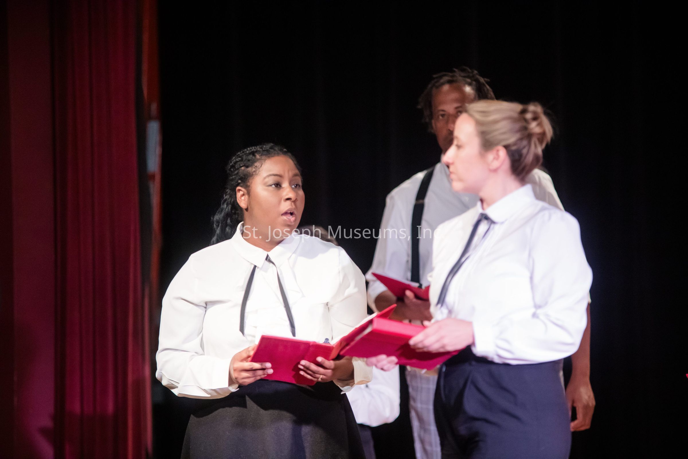 The image depicts a scene on stage with three individuals engaged in what appears to be a theatrical or musical performance. Two people in the foreground, wearing white shirts, black ties, and dark skirts, are holding red booklets, possibly scripts or music, and appear to be singing or speaking to each other. The person on the left has dark hair pulled back, while the person on the right has light hair tied in a bun. In the background, a third individual, partially obscured, is standing and also holding a red booklet, dressed similarly with suspenders. They are positioned against a dark backdrop with red curtains framing the stage, indicating an intimate performance setting. The lighting highlights the concentration and interaction between the performers, creating a focused and expressive atmosphere that suggests a serious or emotionally intense moment in the performance.
