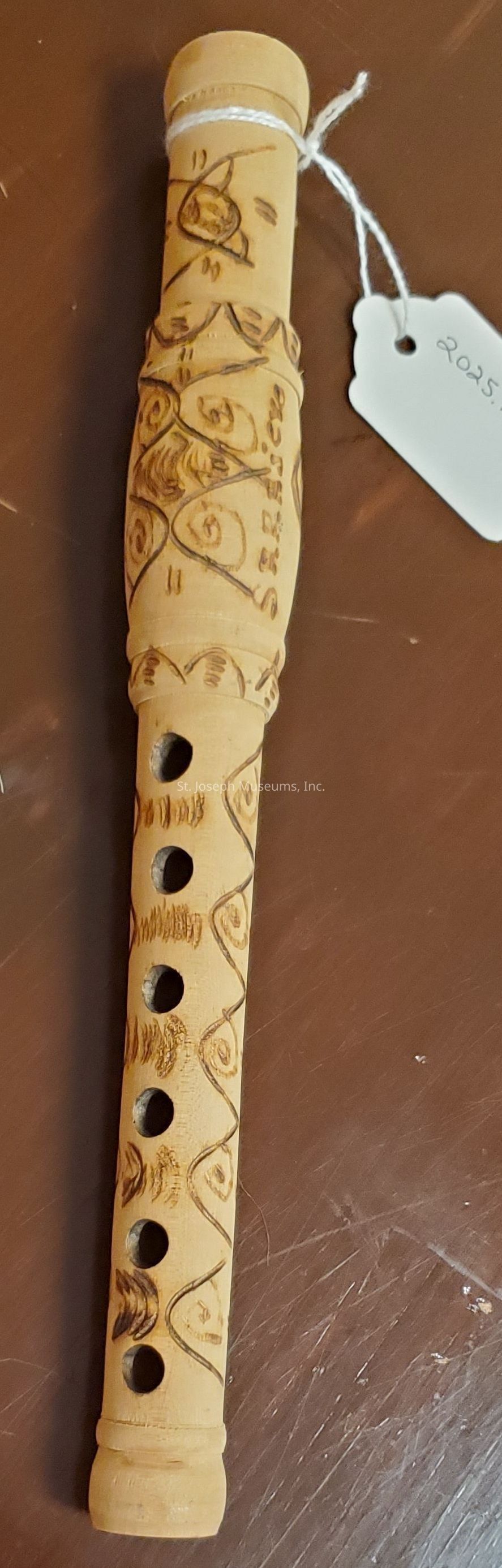 The image depicts a wooden flute with intricate carvings that feature decorative patterns along its body, including swirls and geometric shapes. A small white tag attached to the instrument, possibly indicating a year, further complements the overall craft. The flute is placed on a textured brown surface that provides a contrasting background, emphasizing the unique details and craftsmanship of the instrument.