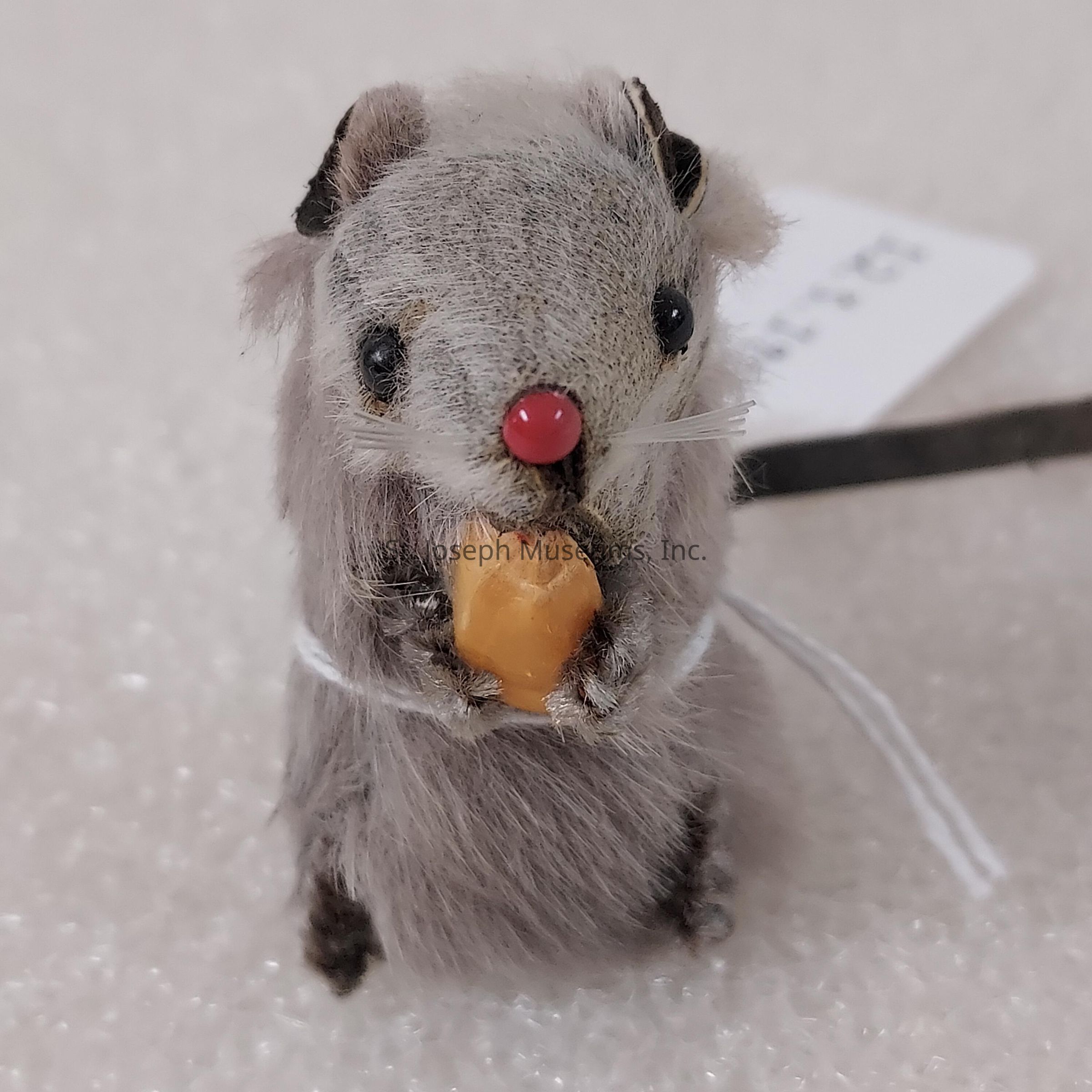The image depicts a small, plush toy mouse with light gray fur and a distinct red nose, holding a small, round object that resembles a piece of food. Its black eyes are prominently set on its face, giving it an alert and curious expression. The mouse is positioned upright, suggesting it is perched or placed on a flat surface. The toy features black ears with slightly darker tips and has a subtle touch of whiskers which add to its detailed craftsmanship. It looks to be tied with a thin white string around its body, adding a playful element to its charming appearance. The background is a neutral, soft-textured surface that complements and contrasts with the mouse, focusing attention on its cute design. A strip of paper with text is visible in the background but its content is not the focal point. The toy appears to be designed for decorative purposes, exuding an endearing quality that makes it suitable for display on shelves or as a whimsical addition to a collection of small plush creatures.