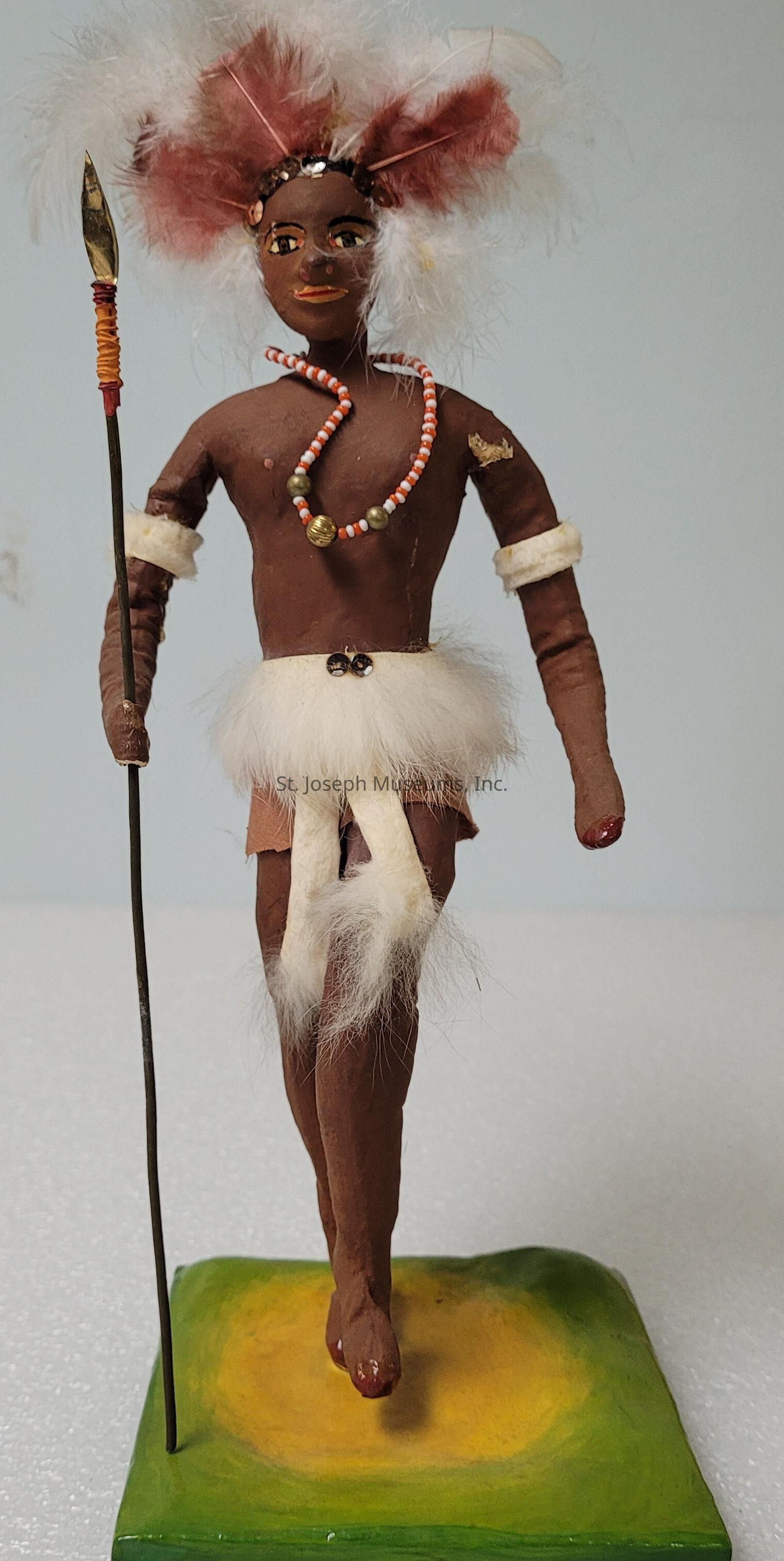 The image depicts a handcrafted figurine of an individual dressed in traditional attire, featuring a deep brown complexion and adorned with a feathered headdress and decorative beads around the neck. The figure is posed walking forward, confidently holding a spear in its right hand. The attire consists of a fluffy white skirt-like garment and additional white fur accents on the arms and legs, adding an element of authenticity and texture. Positioned on a green and yellow base, the figure's expression is detailed with orange accents on the lips, enhancing its vibrant presence. The craftsmanship reflects cultural artistry and attention to detail, with a focus on traditional warrior or ceremonial symbolism.