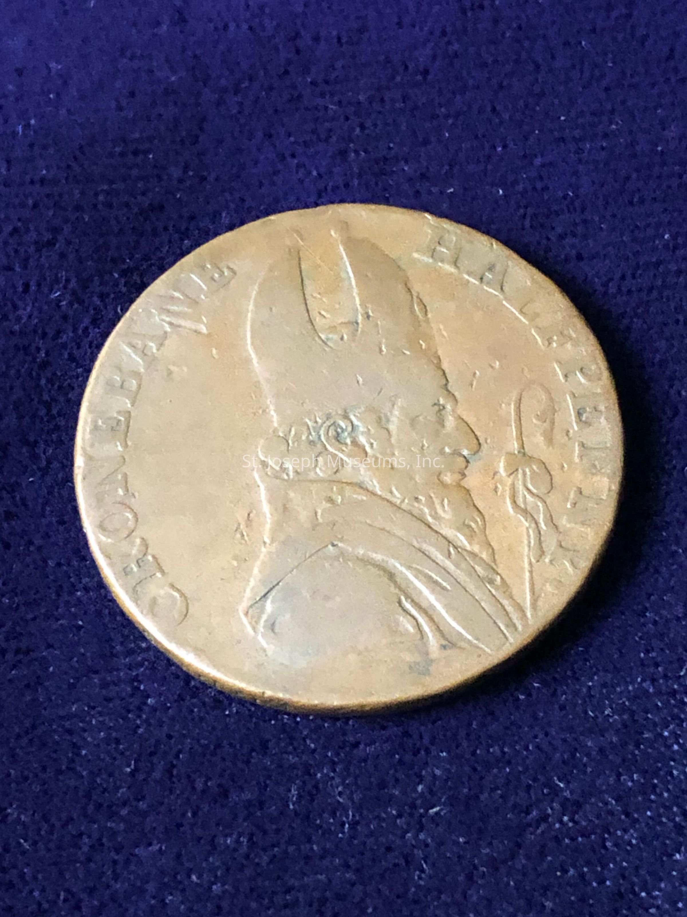 The coin, which appears to be old, features the profile of a figure wearing a distinctive headdress, possibly a bishop’s mitre or similar ceremonial headgear, suggesting ecclesiastical or religious significance. The borders of the coin include an inscription, although some parts are worn and difficult to decipher. The coin has a golden hue, indicative of tarnish or age, and its surface shows signs of wear and scratches, hinting at a long history. The overall composition suggests that the coin may have both numismatic and historical value.