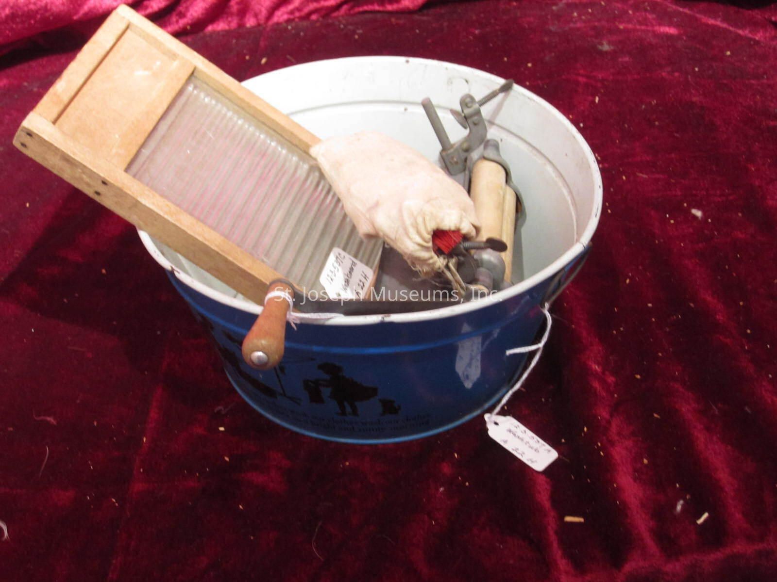 12.3.537 A-c: The image shows a vintage laundry setup placed on a rich red fabric background. A galvanized metal wash tub, painted blue with an illustration of a person doing laundry, is filled with an assortment of old-fashioned laundry tools. Leaning against the tub's edge is a wooden washboard with a ridged, clear surface used for scrubbing clothes. Inside the tub, there's a manual clothes wringer with wooden rollers and metal clamps, which would typically be used to squeeze excess water from garments after washing. A small cloth bag with a drawstring lies inside, possibly containing soap or a similar washing aid. Each item bears a tag with handwritten prices, suggesting they might be for sale in an antique shop or market. The items evoke a nostalgic glimpse into past domestic chores, showcasing the traditional methods of washing clothes before the advent of modern washing machines. This setup highlights both the labor-intensive nature of household tasks in the past and the craftsmanship of vintage home appliances.