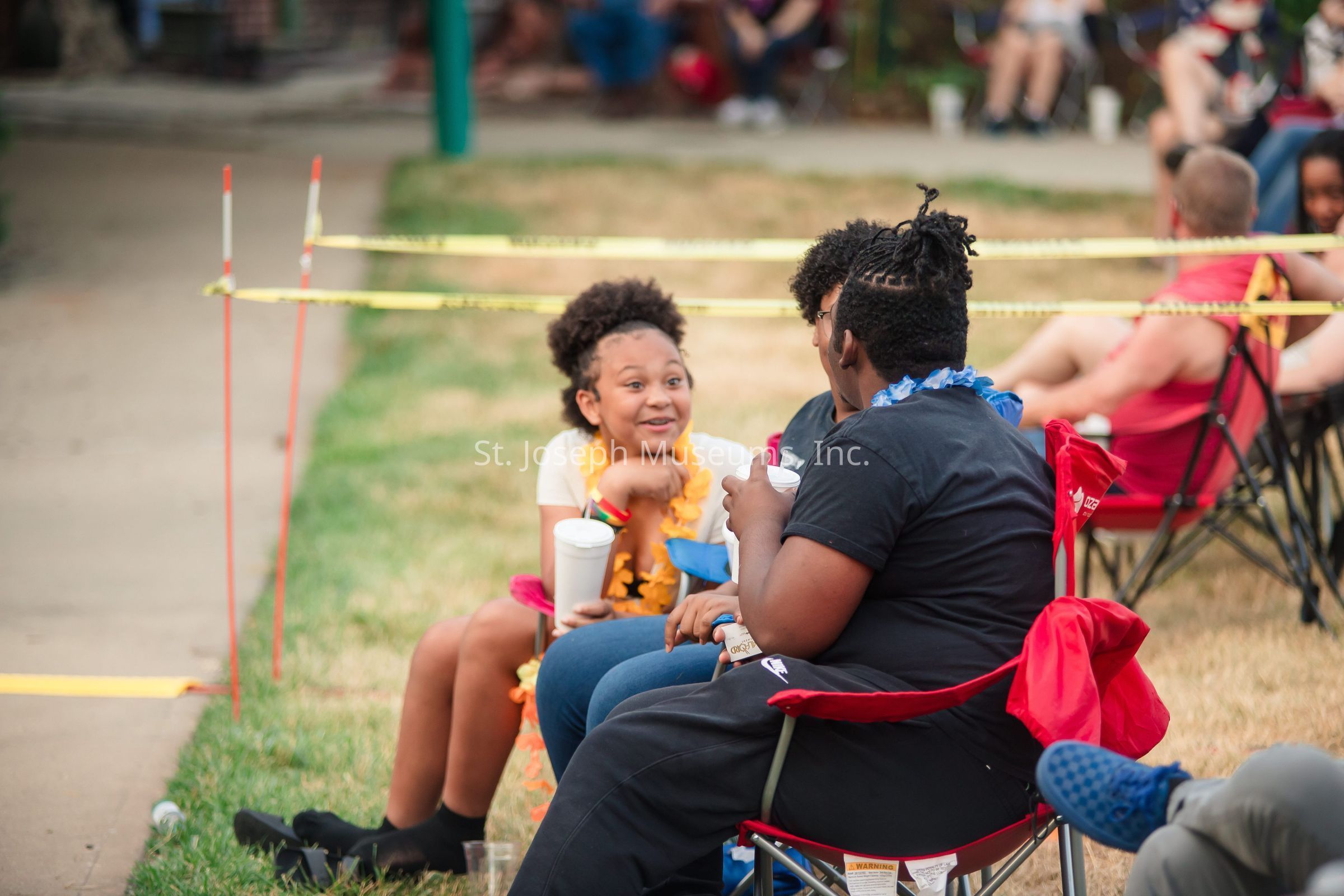 In the image, a group of people wearing leis are sitting on red chairs and enjoying each other's company in an outdoor setting. One young girl, who has a joyful expression, is holding a large cup and engaging in conversation with the others around her. The atmosphere appears relaxed and festive, suggesting some sort of gathering or event. The ground is grassy, and the area is partially surrounded by caution tape, while more people can be seen seated in the distance against a paved walkway.