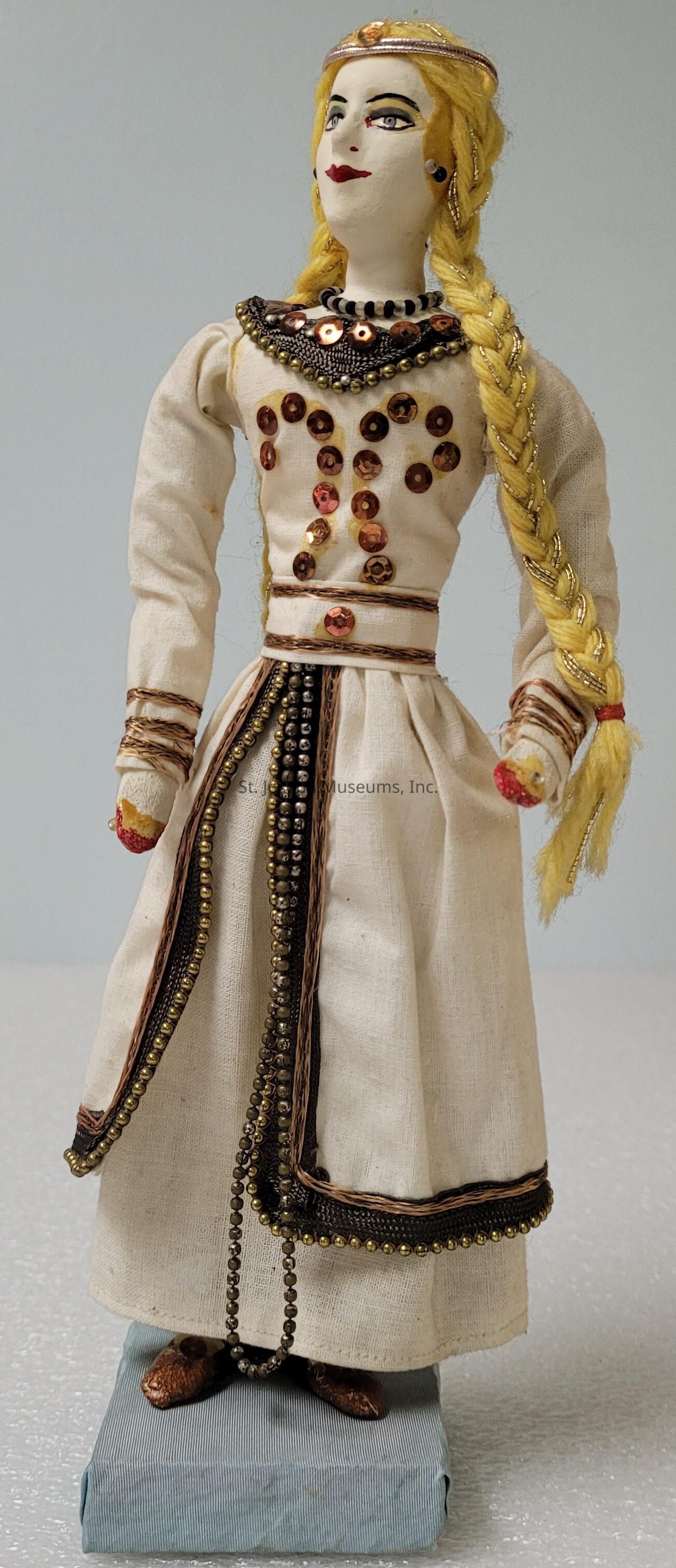 The image showcases a handmade doll featuring a detailed design with a traditional, richly decorated garment. The doll stands on a small platform, wearing a long off-white dress adorned with intricately sewn sequins and beadwork in gold and bronze tones. The doll's dress is complemented by braided gold-tone accents and matching trim around the neckline and waist, enhancing its elegant appearance. This doll has striking facial features, with painted deep red lips and dark eye makeup, giving it a distinct personality. Its hair, crafted from yellow yarn, is neatly braided to one side, adding a touch of rustic charm. This beautifully crafted piece is a testament to intricate artistry, evoking cultural and historical aesthetics through its attire and overall design. The attention to detail, from the stitching patterns to the placement of decorations, highlights the craftsmanship involved in making such a unique item. The use of various textures and materials not only represents skillful technique but also emphasizes the richness and allure of traditional attire, making it a fascinating subject.
