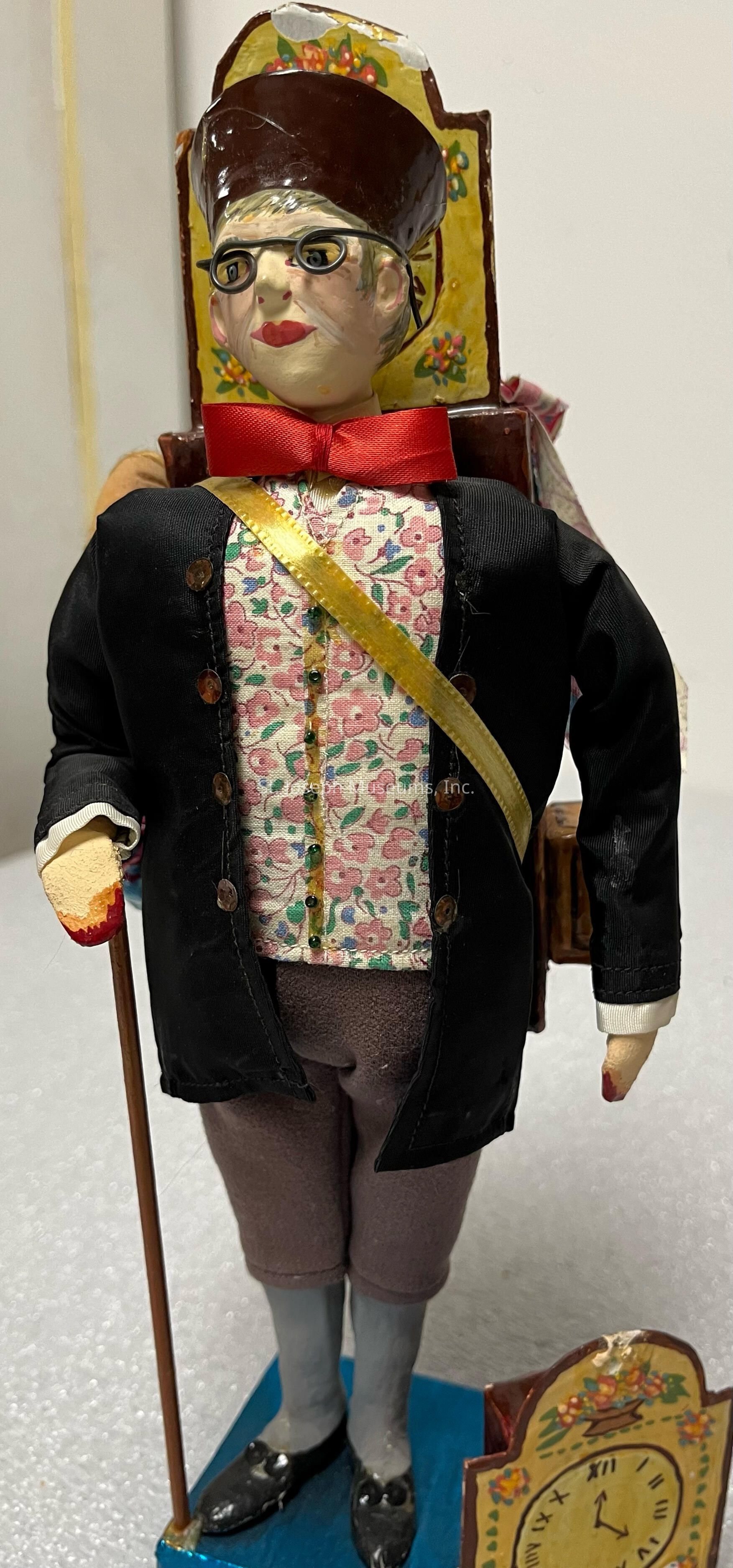 The image shows a handcrafted figurine that resembles a traditional European clockmaker. It is dressed in a black jacket adorned with a floral-patterned vest and a striking red bow tie. The figure wears glasses and a dark hat, carrying a detailed clock on its back with vibrant floral designs. Its right hand holds a wooden cane, adding to the classic appearance. The figurine stands on a blue base, with a distinctly painted clock face in front, featuring Roman numerals and small floral decorations. The craftsmanship reflects a charming blend of whimsical art and cultural storytelling.