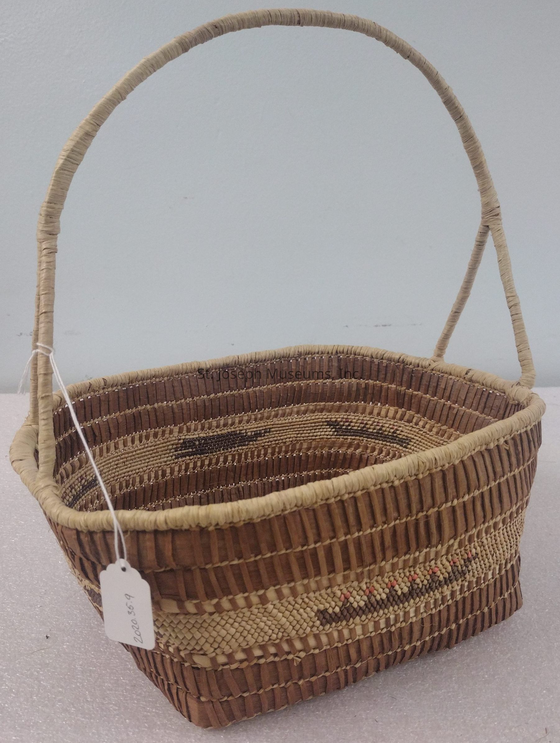 The image shows an intricately woven basket featuring an arching handle made from natural fibers. The basket has a rectangular shape with a sturdy construction that highlights the artisan's skill. Its design includes a pattern in earthy tones of beige, brown, and a hint of red. The surface is adorned with alternating bands of tight and loose weaving, providing textural contrast and visual interest. The basket is positioned on a textured, light surface, set against a plain light blue wall background, suggesting it is on display, likely in a gallery or store setting, where its craftsmanship and quaint design can be appreciated fully.
