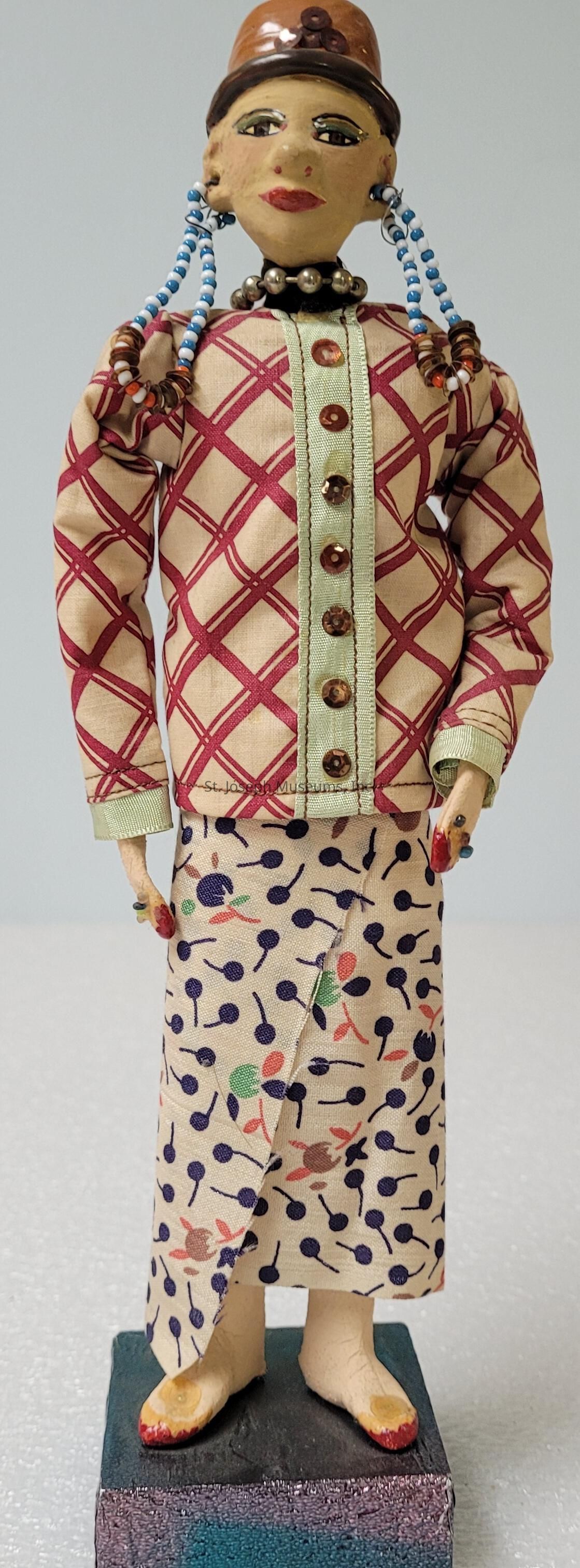 The image depicts a handcrafted doll adorned in a colorful and intricate outfit. The doll wears a patterned blouse with a crisscross red and beige design, accented with a strip of green fabric featuring brown buttons down the front. The skirt displays a pattern of multicolored, abstract shapes resembling cherries on a cream background. The doll’s facial features are carefully painted, with expressive eyes and red lips. Its hair is adorned with beads, and it wears a hat on top. The doll stands on a small wooden platform, highlighting its detailed and artistic craftsmanship.