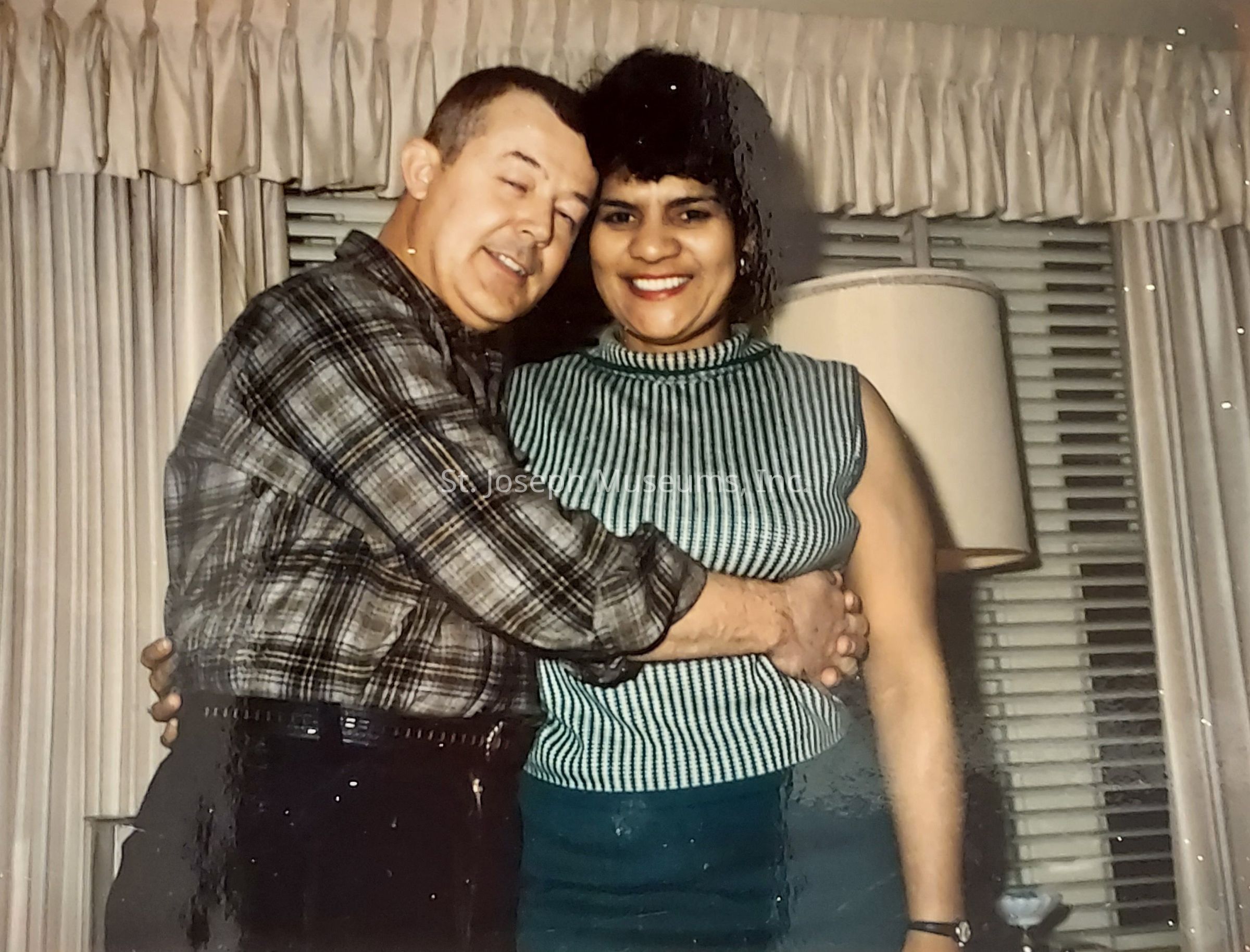 In this vintage photograph, a man and woman are warmly embracing each other while standing indoors, suggesting a sense of affection and closeness. The man is wearing a plaid shirt and has a gentle smile, while the woman, wearing a green and white striped sleeveless top, is smiling brightly. Behind them, the room is decorated with neutral curtains and a lamp, adding a cozy and intimate atmosphere to the scene. The overall mood of the photo is joyful and nostalgic, capturing a tender moment between the two people.