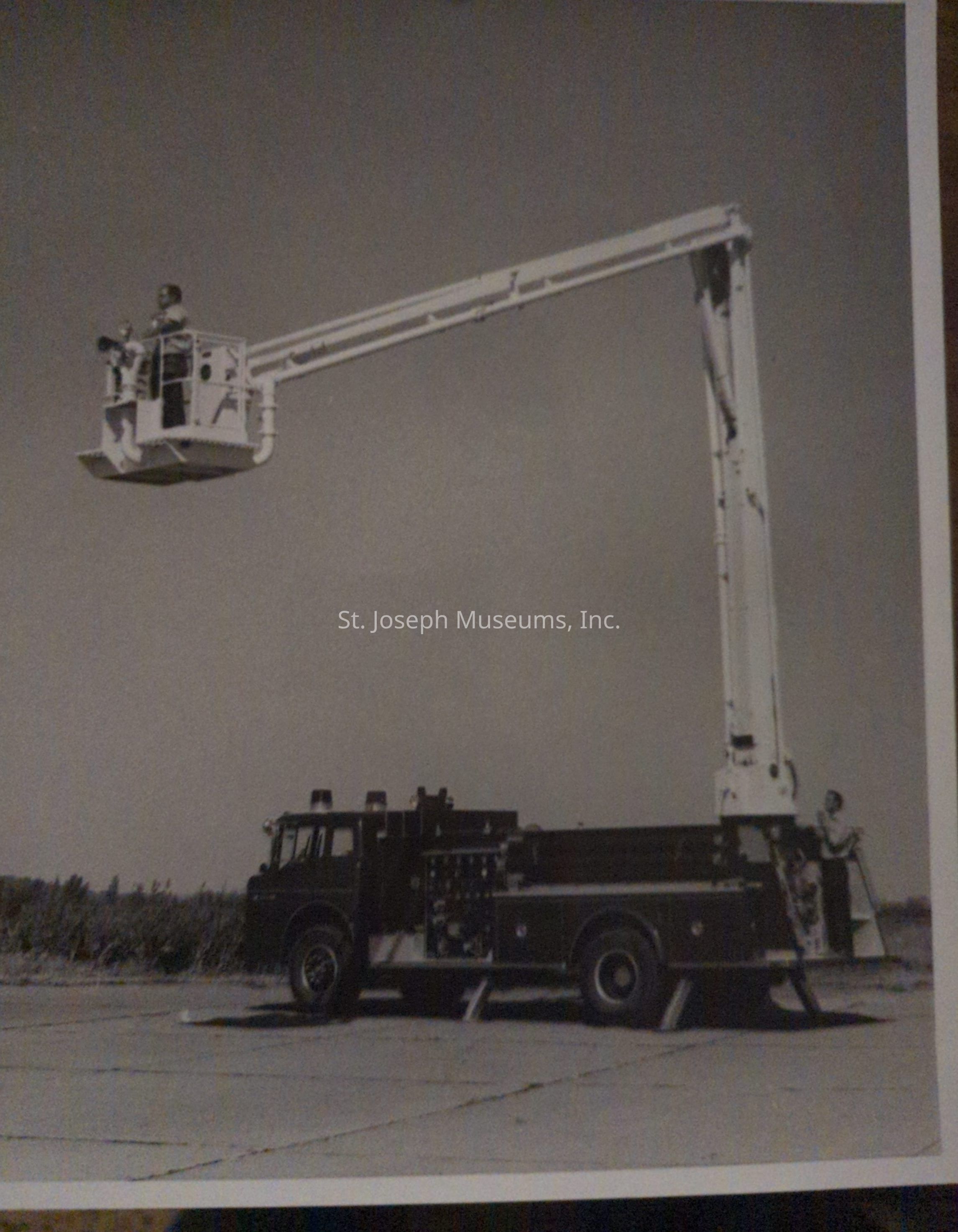 Black-and-white photograph depicting a fire truck from Snorkel Co., positioned on Lake Road and Arizona, featuring an extended aerial platform. A person operates the platform high above, showcasing its impressive reach and functionality, with the truck placed on a flat road, illustrating its readiness for firefighting operations.