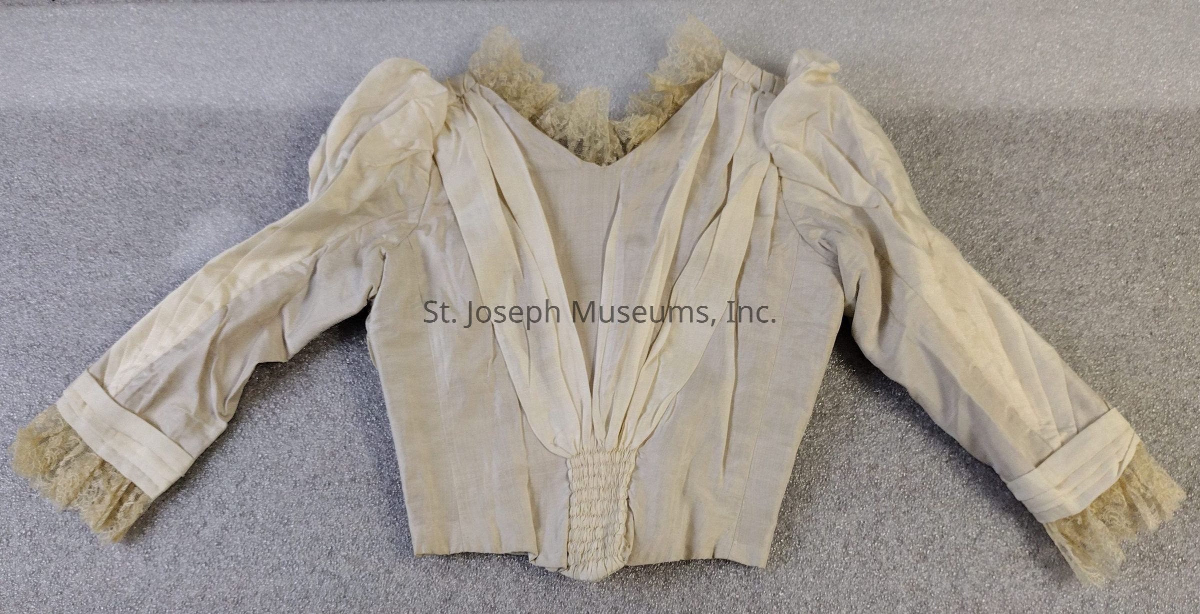 The image showcases a wedding dress bodice with intricate detailing. The fabric appears to be a light, possibly ivory material with gathered pleats creating a V-shaped design at the front, leading to a ruched section at the waist. The long sleeves are gathered at the shoulders, with delicate lace trimming at the cuffs and neckline.