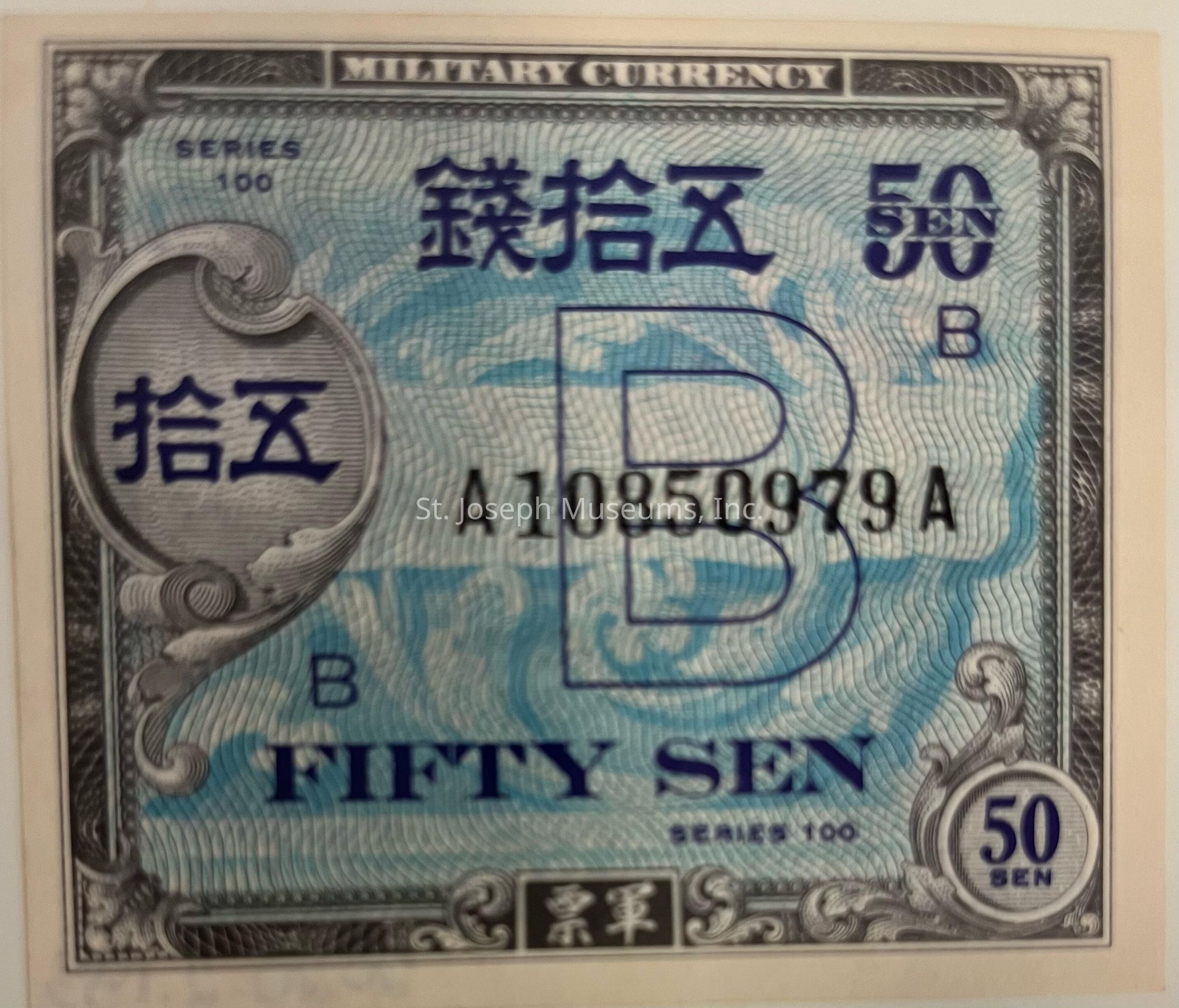 The image displays a piece of military currency, specifically a 50 sen note from Series 100. The design prominently features intricate patterns in shades of blue and black, with the denomination "FIFTY SEN" clearly printed in large font across the lower portion. The top of the note is inscribed with "MILITARY CURRENCY," and a prominent letter "B" is visible in the center. Additionally, the note includes a serial number "A10850979A" printed in black. Ornamental designs frame the corners and edges, and there is an oval section on the left containing foreign text, likely indicating value. The number "50" is repeated in a circular design at the lower right. The note exhibits a classic, historical aesthetic typical of military-issued monetary items.