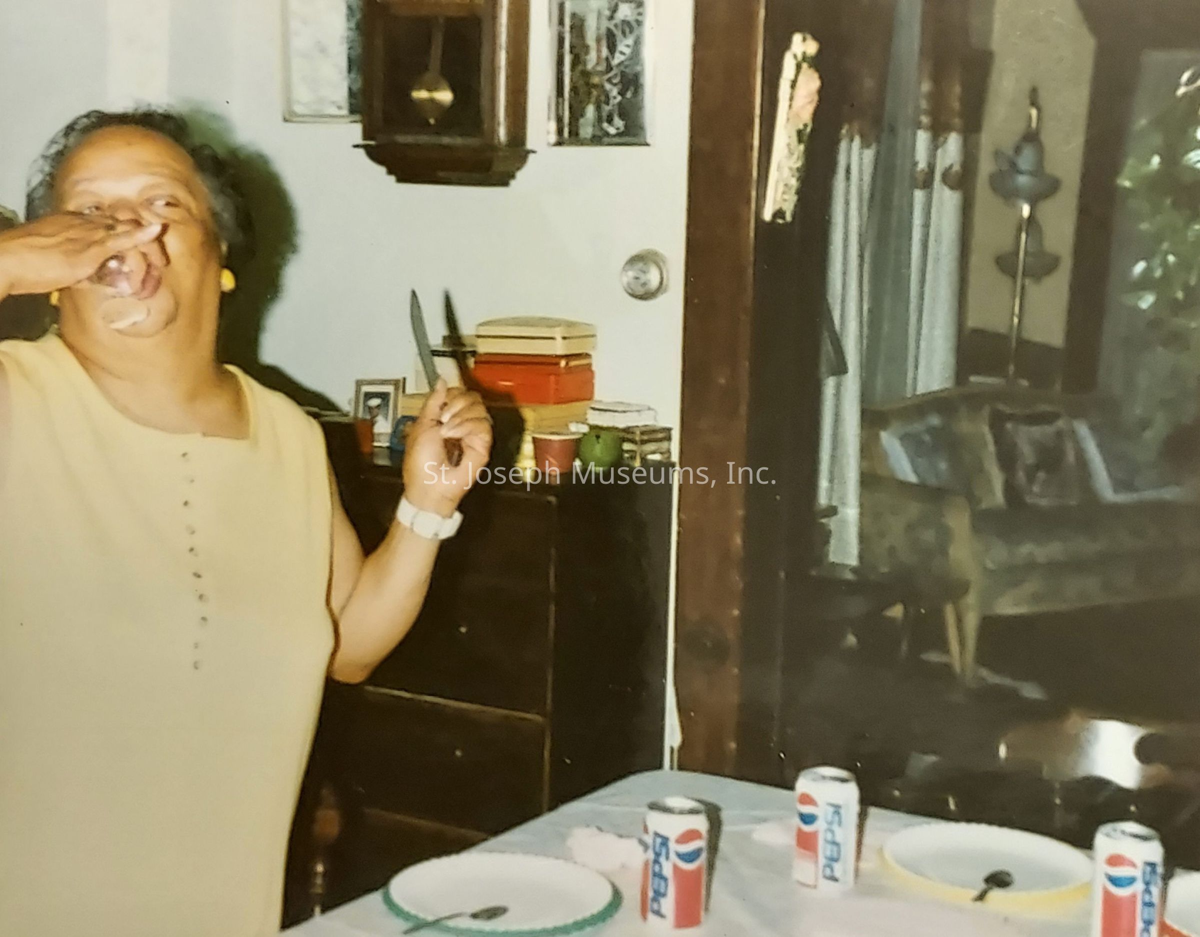 The image captures a joyful moment where a person is standing in a room holding a knife with one hand while wiping their mouth with the other, suggesting they might be enjoying a meal or snack. On the table before them are several Pepsi cans and plates, indicating a gathering or celebration. The person wears a sleeveless top, yellow earrings, and a watch, exuding a relaxed and casual vibe. In the background, a cozy living space is visible, with an upholstered chair, a decorative floor lamp, and patterned curtains adding to the inviting atmosphere. The scene conveys a sense of warmth and conviviality.