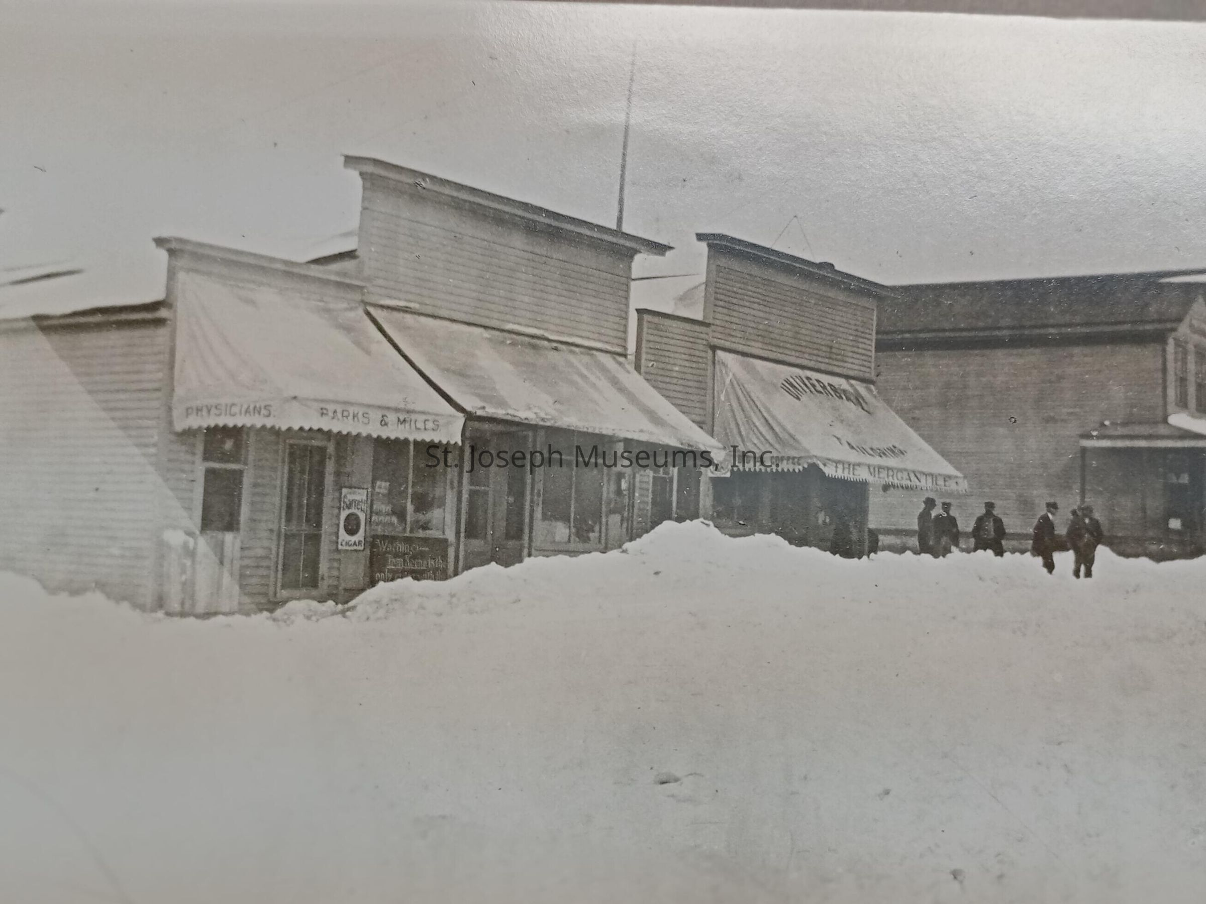 Black-and-white image depicting a row of old-fashioned, western wooden buildings with awnings, including a physician's office named "Parks & Mills" and a mercantile store; the ground is covered with a thick layer of snow and a group of people, dressed in dark, heavy coats, are seen walking in the distance near the storefronts.