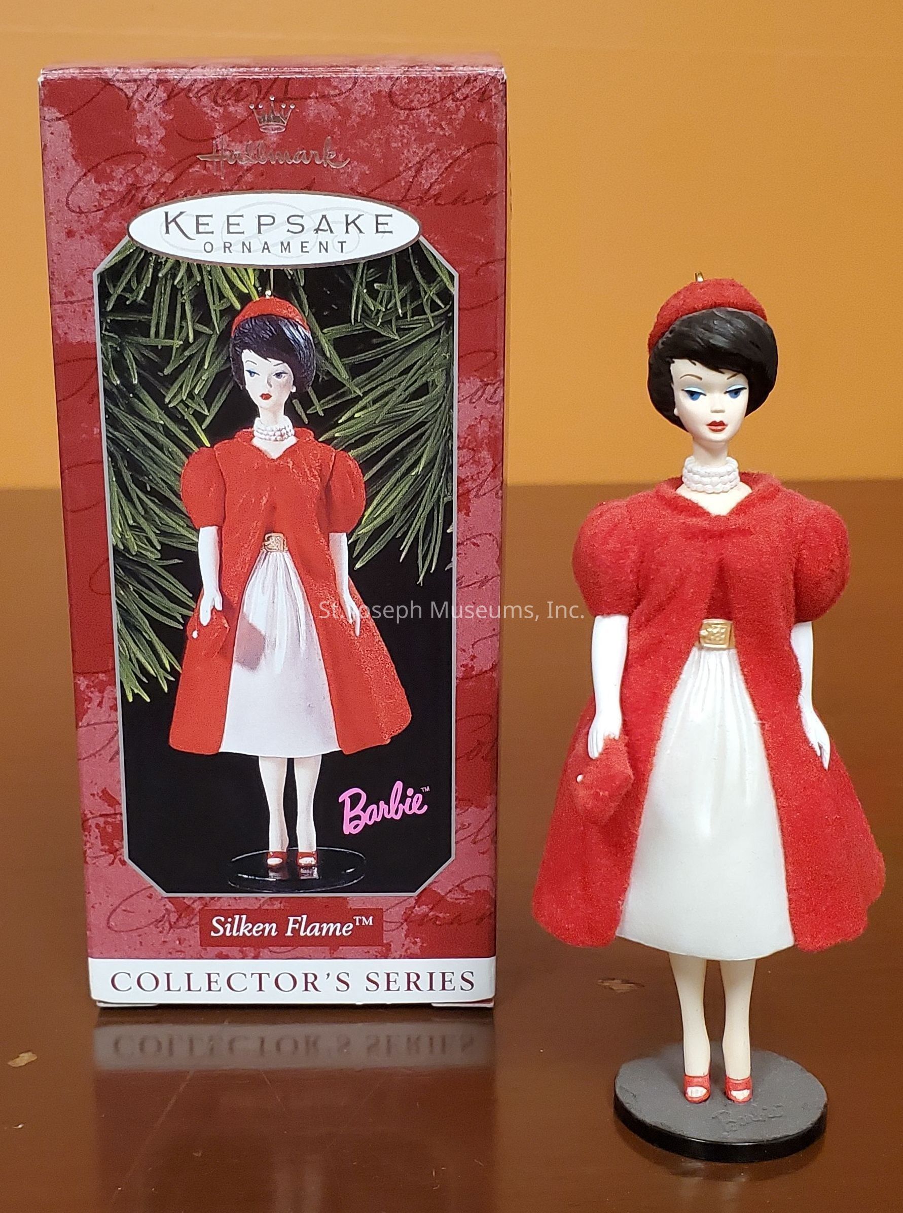The image shows a Hallmark Keepsake Ornament from the Collector's Series featuring a vintage-inspired doll dressed in a classic ensemble. The doll is styled with a black bob hairstyle and wears a striking red coat over a white dress, paired with a matching red hat and shoes. The packaging showcases the same image of the doll, emphasizing its vibrant colors against a festive green background. The text on the box reads "Silken Flame™" and "Barbie™," indicating this is a collectible item from a well-known series. The overall presentation evokes a nostalgic, elegant charm, characteristic of mid-century fashion.