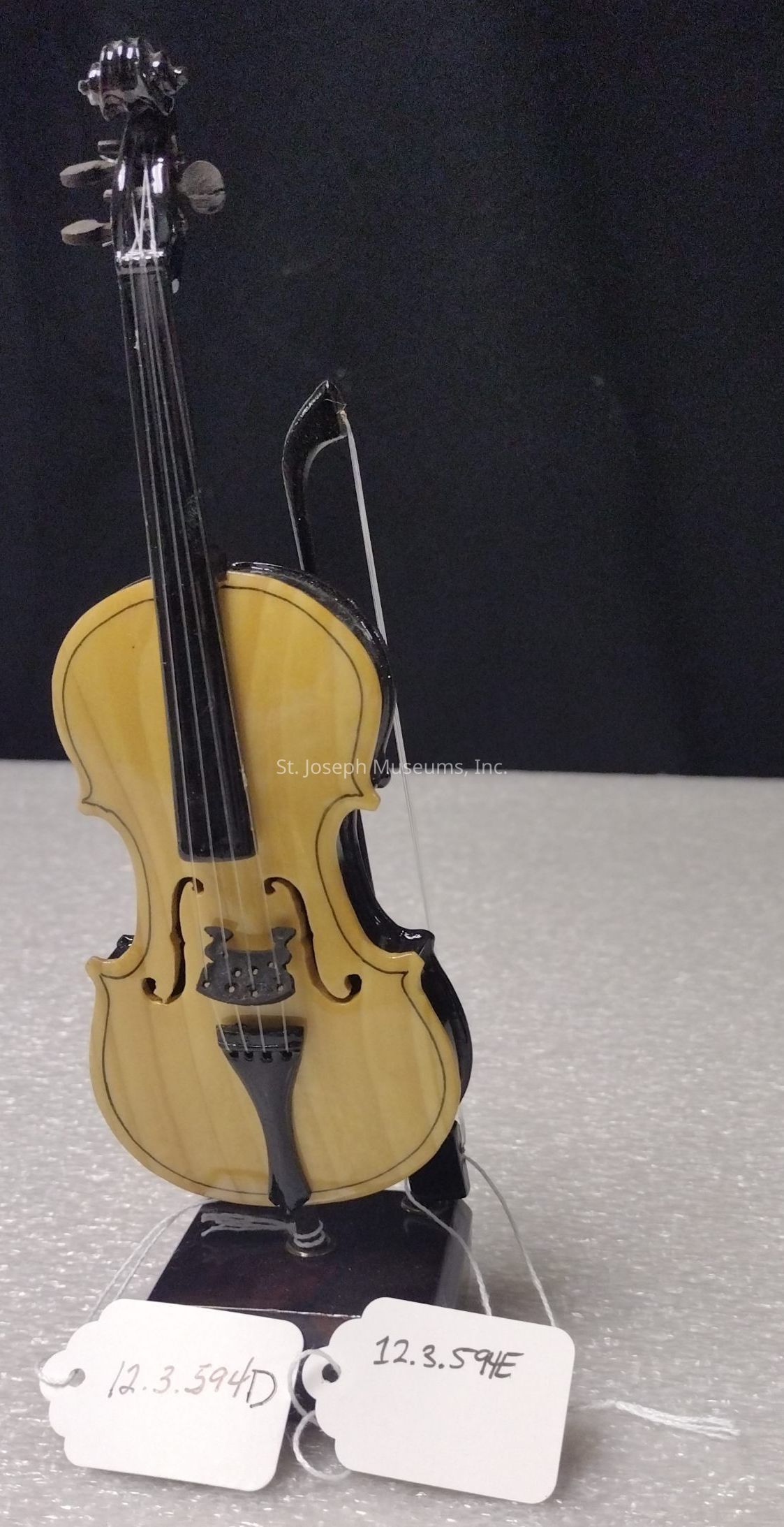 The image depicts a small, intricately crafted violin model resting upright on a textured surface, with the body of the violin showcasing a light wood grain finish and elegantly curved black detailing on the edges; it has four strings stretching from the neck to the tailpiece, and is accompanied by two white tags that have numbers written on them, all set against a dark, neutral background which helps accentuate the fine details and craftsmanship of the miniature instrument, emphasizing its aesthetic appeal and delicate construction as a representation of a classic violin.