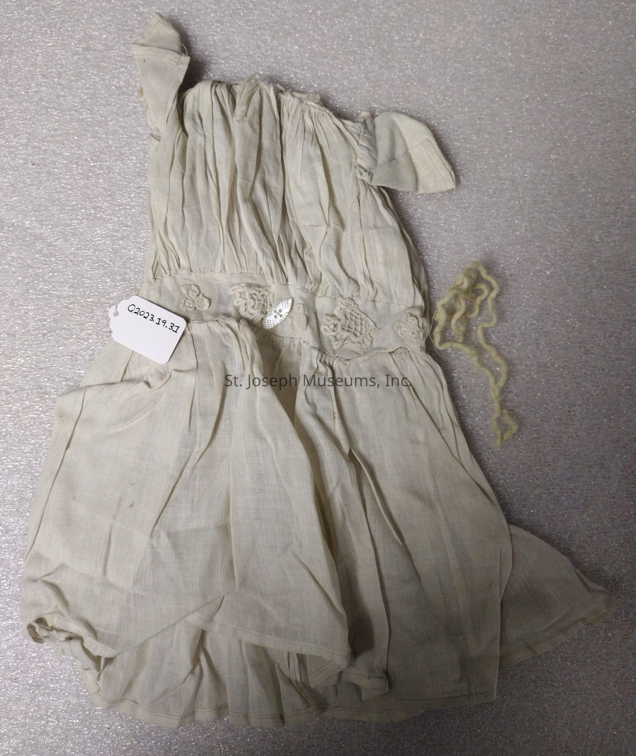 The image shows a small, vintage-looking garment, possibly a child's dress, made from a light, cream-colored fabric with a fine, delicate texture. The dress is laid out on a textured, gray surface, and it features intricate details such as pleating and lace embellishments along the waistline. A tag with the inscription "Q2023.19.32" is attached to the dress, suggesting it could be an item cataloged for historical reference or archival purposes. Beside the dress, there is a small piece of yellowish, twisted material, possibly a fragment of thread or yarn, lying casually on the surface. The dress appears to be in fragile condition, with slight creases and an overall antique appearance, indicating it may be of considerable age or historical significance.
