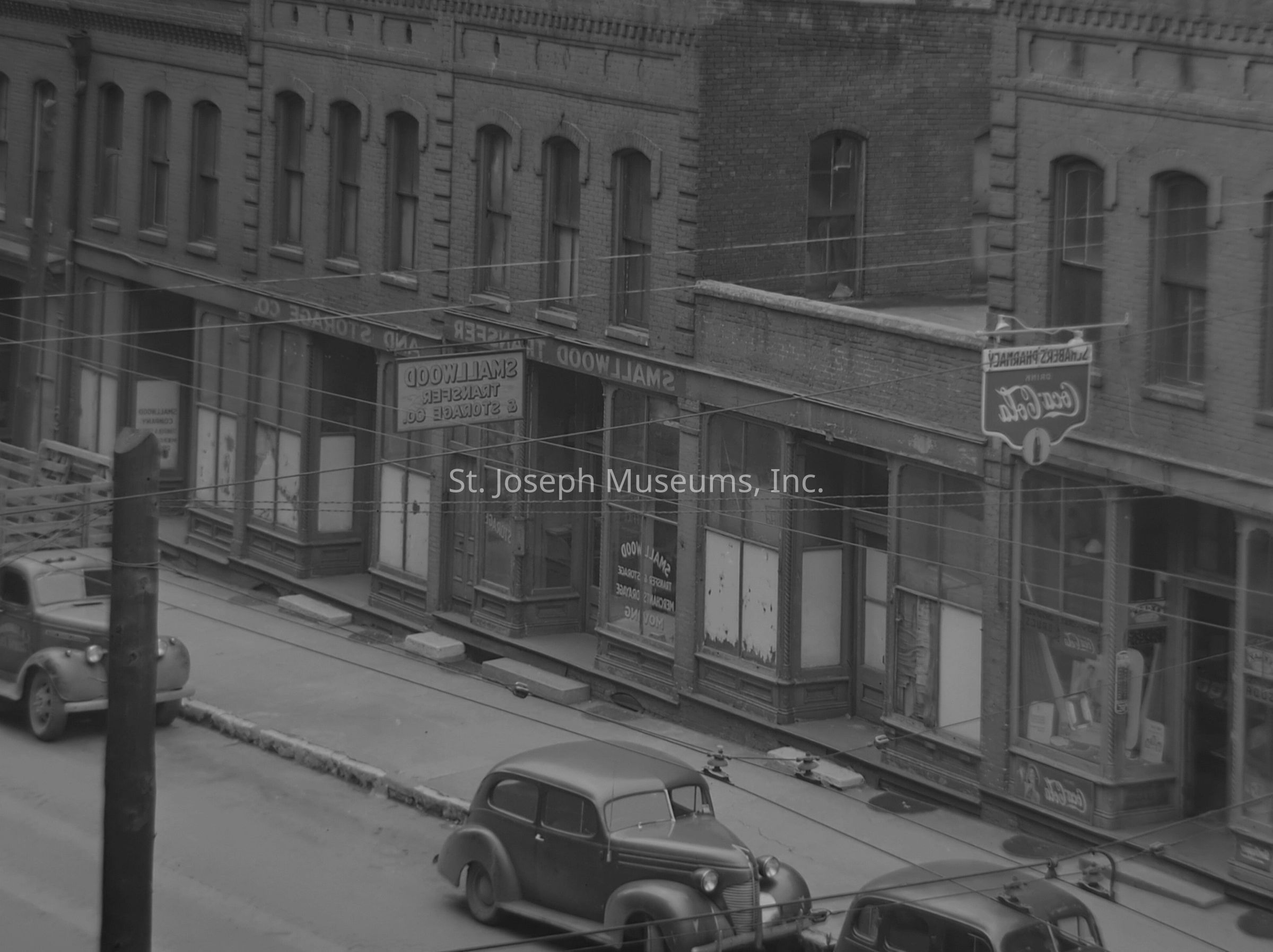 Black-and-white image depicts a historical street view of Smallwood Transfer & Storage Co. at 301 S. 3rd St. in St. Joseph, Missouri, showcasing a row of old brick buildings with large window displays. Automobiles are parked along the curb, while overhead electric cables crisscross the scene. A weathered Coca-Cola sign hangs above one of the storefronts. The windows of the buildings bear signs of wear.