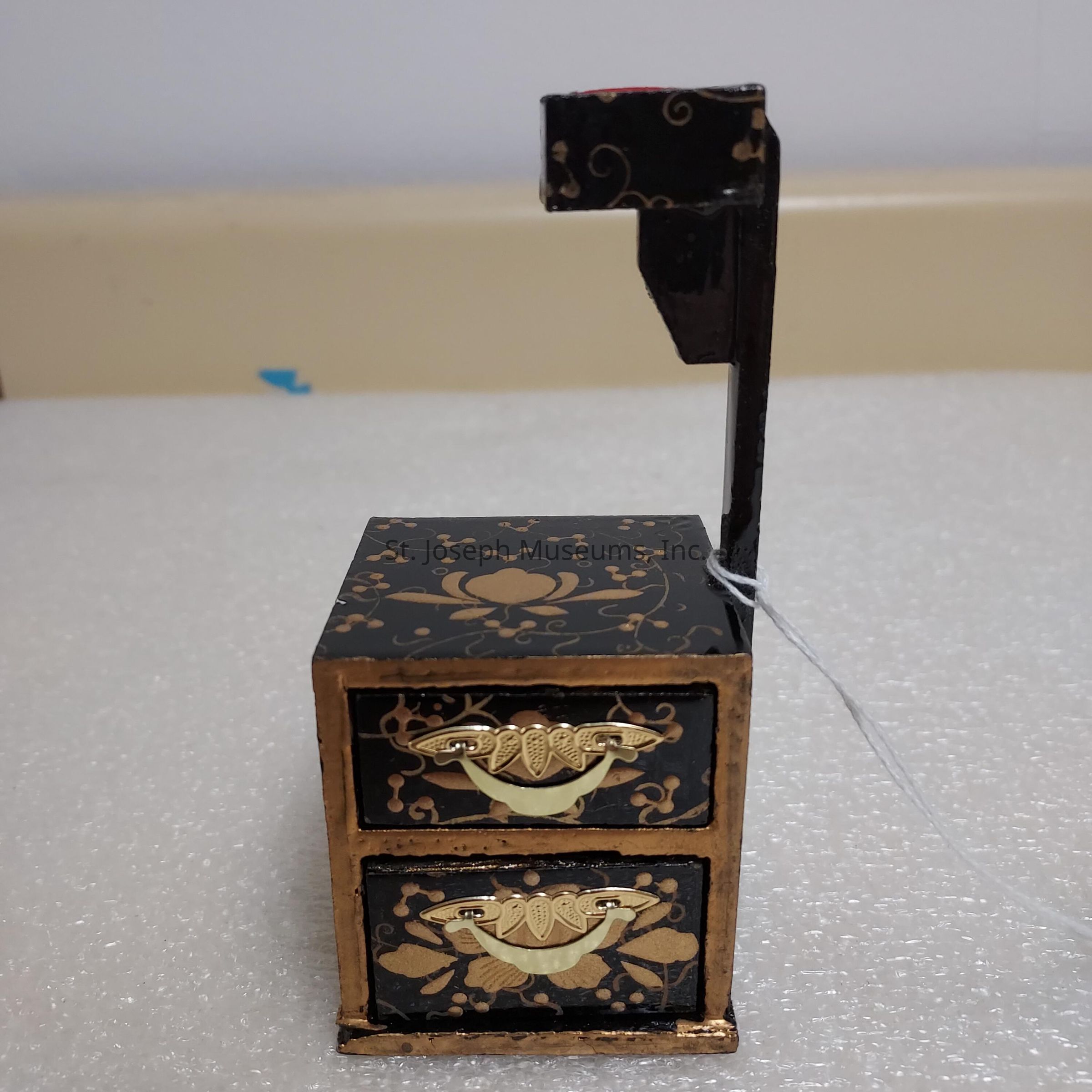 The image features a small, ornate black lacquered box, possibly a miniature tansu or decorative chest, with intricate gold patterns adorning its surface, including floral and vine motifs. The box has two drawers, each featuring elegant, gold-colored handles shaped like stylized leaves or fans, contributing to its artistic appearance. A vertical, angular structure extends from the top of the box, possibly serving a functional or decorative purpose. The entire piece rests on a textured, white surface and exudes a sense of traditional craftsmanship, likely indicative of East Asian, particularly Japanese, design aesthetics.
