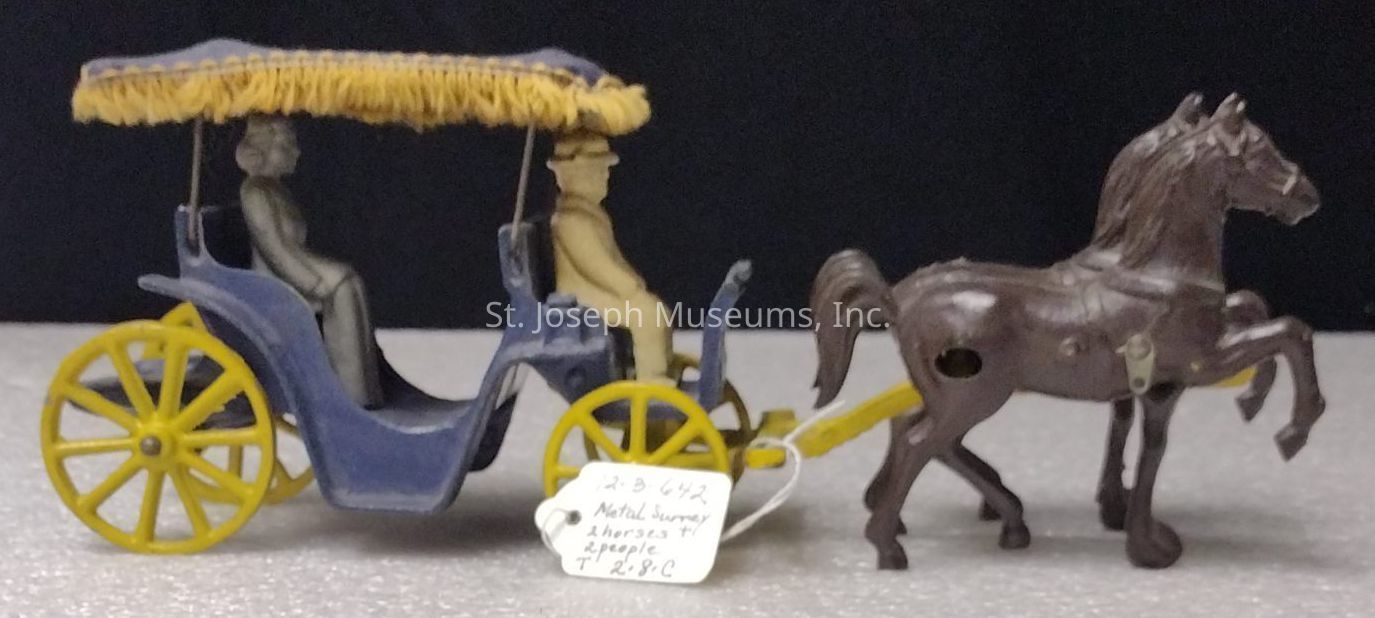 The image shows a vintage metal toy set featuring a horse-drawn carriage. The horse, crafted in brown, is in motion with one front leg raised, suggesting movement. Attached to the horse is a blue carriage with bright yellow wheels and a fringed yellow canopy. Inside the carriage sit two figures, one in gray and the other in yellow, styled in a manner reminiscent of early transportation. A small white tag hangs from the carriage, with handwritten text indicating a catalogue number and description, adding an antique or collectible quality to the piece, and suggesting its place in a collection or display.