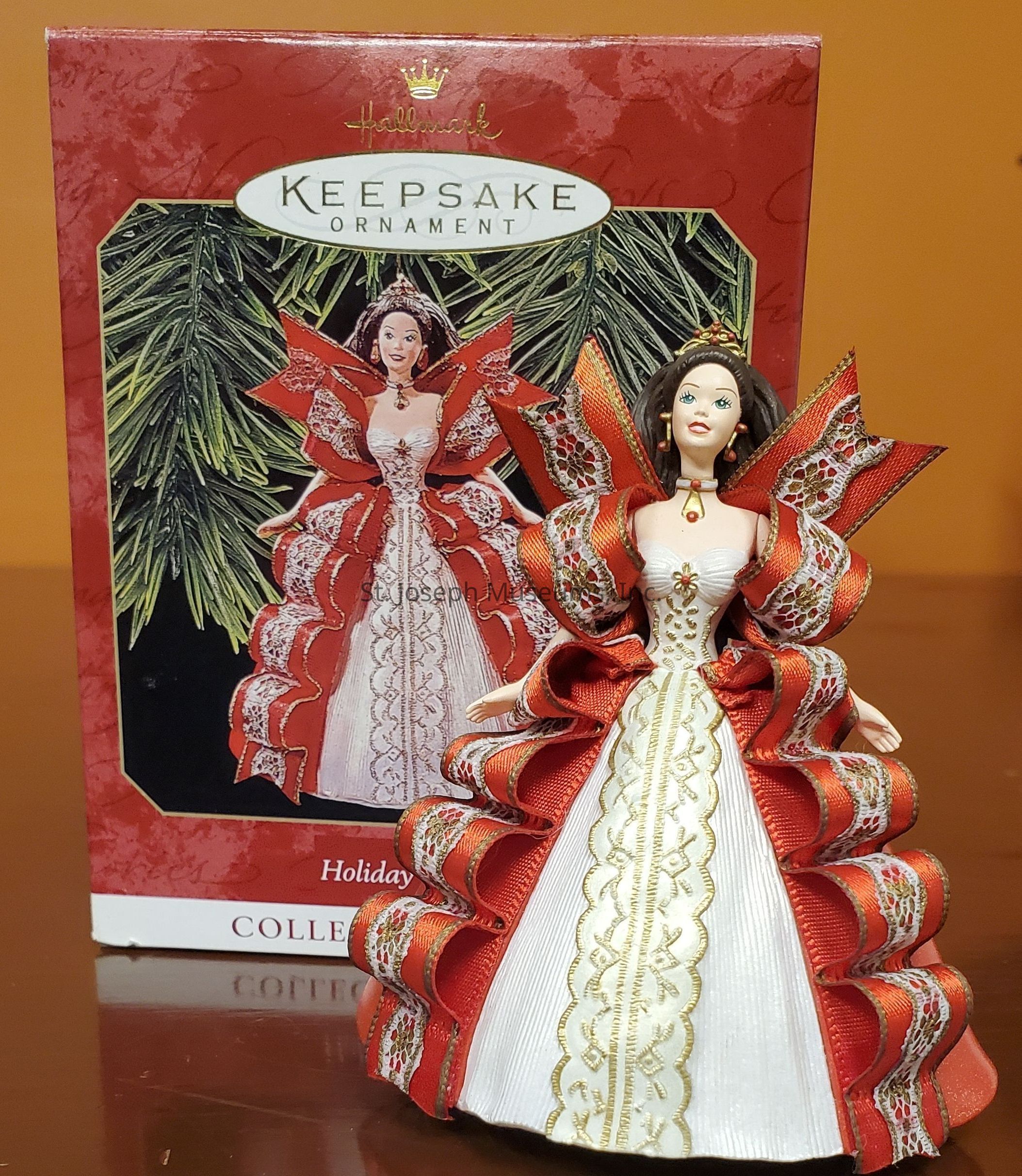 The image features a Hallmark Keepsake Ornament displayed in front of its ornate packaging. The ornament is a detailed figurine of a woman dressed in an elaborate gown made up of vibrant red and white colors, accented with gold and lace details. The dress is designed with layered, ruffled sleeves and a structured, elegant skirt that cascades smoothly. The woman in the ornament is depicted with a composed expression, wearing a crown and jewelry that complement her attire. The packaging behind echoes the ornament’s design, showcasing an illustration of the same figure set against a background of lush, green holiday foliage. The words "Keepsake Ornament" are prominently displayed on the packaging, underlining its status as a collectible item. The overall presentation of the ornament and its packaging exudes a festive holiday spirit, likely intended for use as a Christmas decoration. The craftsmanship highlights the intricate detailing typical of collectible items, making it a charming addition to any holiday decor.