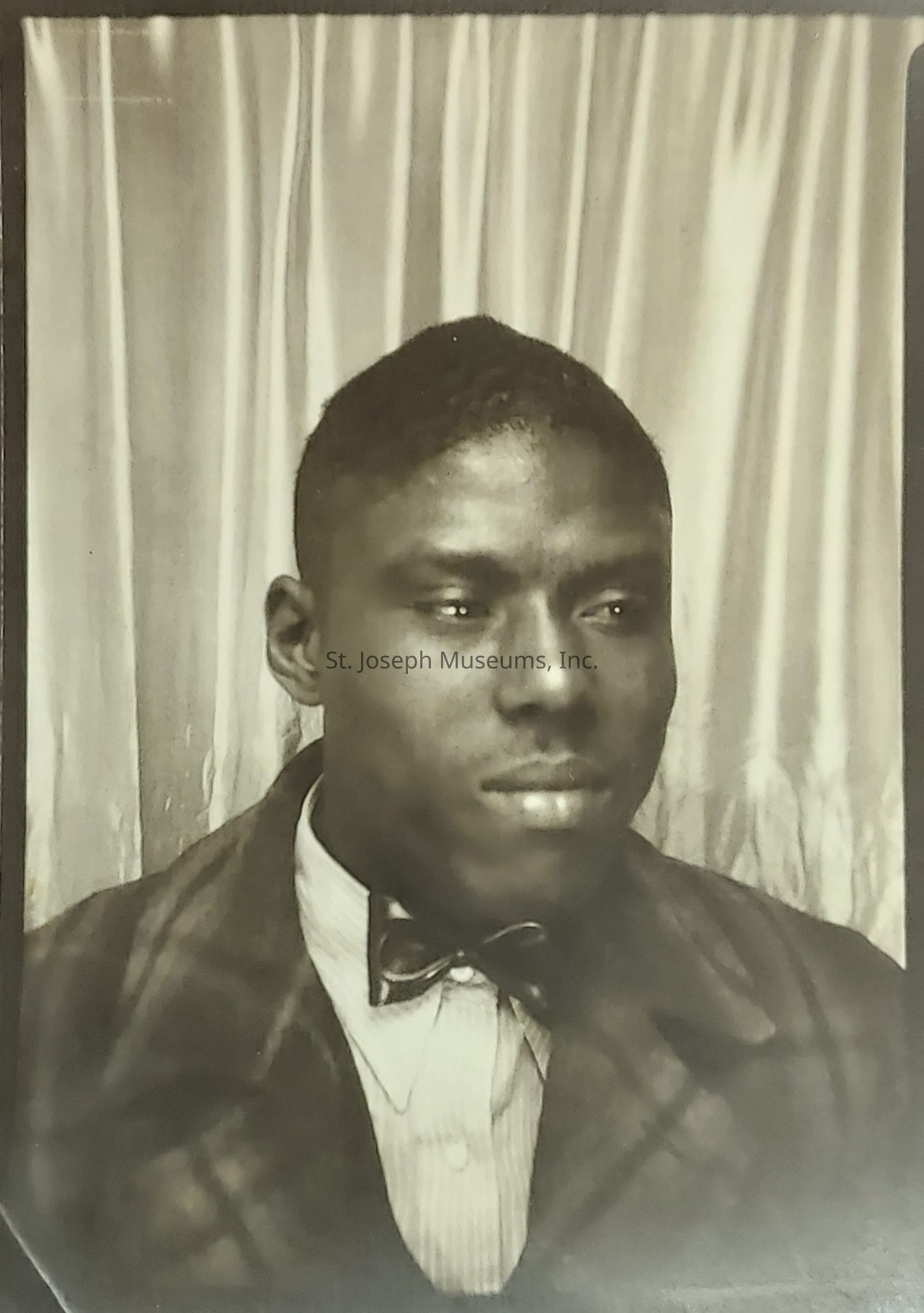 The sepia-toned photograph captures a resolute and dignified man in a vintage setting, framed by a backdrop of draped curtains that evoke a sense of timelessness; his attire, which includes a checked coat over a striped shirt and a neatly tied bow tie, suggests a formal occasion or period during which formality was a prominent feature of everyday apparel—he faces the camera slightly turned, his eyes gazing thoughtfully into the distance, reflecting a sense of introspection and calm confidence, while the meticulous grooming of his short hair completes his polished appearance, adding to the visual narrative of a bygone era characterized by elegance and restraint, and thus the image, with its smooth textures and soft shadows, not only offers a glimpse into the fashion and aesthetics of a previous generation but also invites viewers to ponder the personal history, thoughts, and aspirations of the individual captured within this moment, preserved as an enduring testament to individuality and the timeless human experience.
