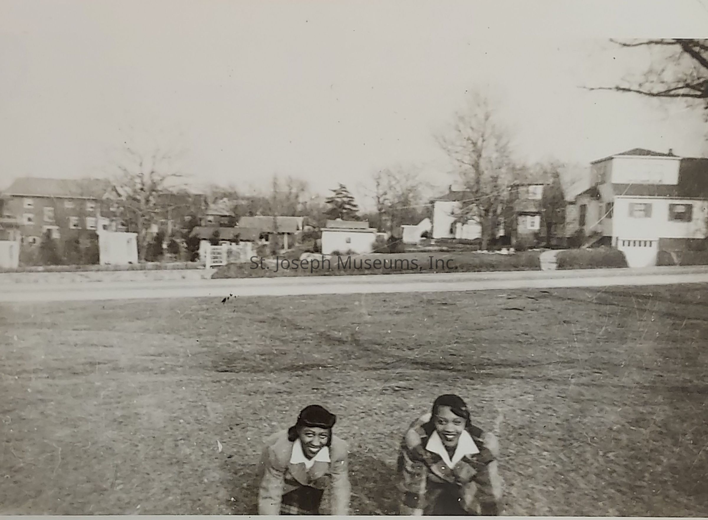 In this black-and-white photograph, two people are crouched down and smiling broadly in an open grassy area with a backdrop of residential neighborhood houses. The landscape is devoid of leaves, indicating a possible fall or winter setting. The individuals are dressed warmly in coats, reflecting the colder season. Behind them, the neighborhood consists of multiple houses, with varying architectural styles, lined along a quiet street. The scene exudes a sense of nostalgia and captures a candid, joyful moment between the two as they enjoy their time outdoors.