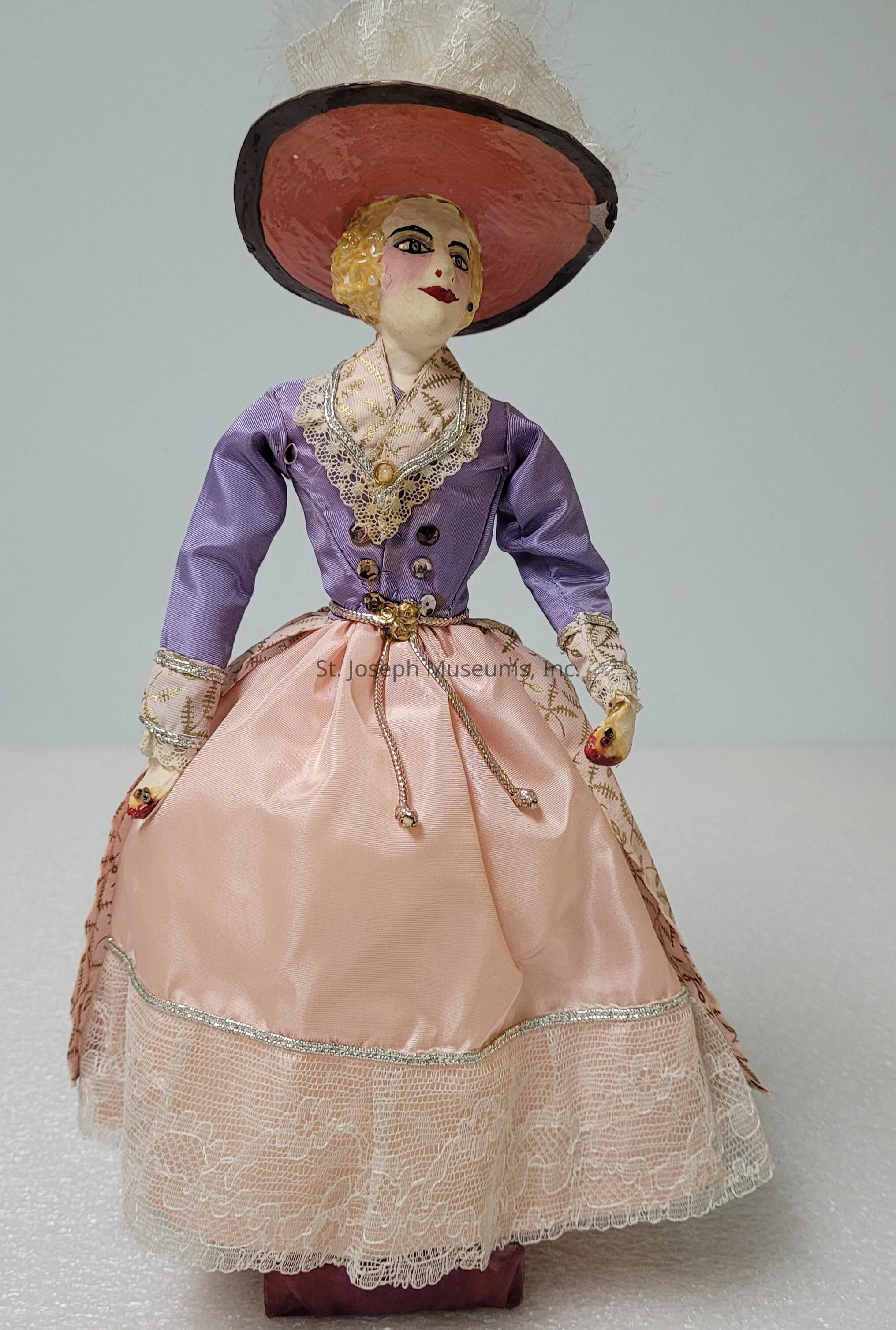 The image showcases an elegantly crafted doll dressed in a vintage-style outfit, featuring a detailed lavender jacket with lace and gold accents, paired with a pink, flowing skirt adorned with a lace hem. The doll's attire is further highlighted by a wide-brimmed hat in pink, topped with delicate fabric embellishments, adding a touch of sophistication. Its face, painted with fine features, includes rosy cheeks and expressive eyes, giving it a lifelike charm. The combination of intricate details and period attire suggests a nostalgia for classic fashion, making the doll a unique and artistic collectible item remarkable for its craftsmanship and attention to detail.