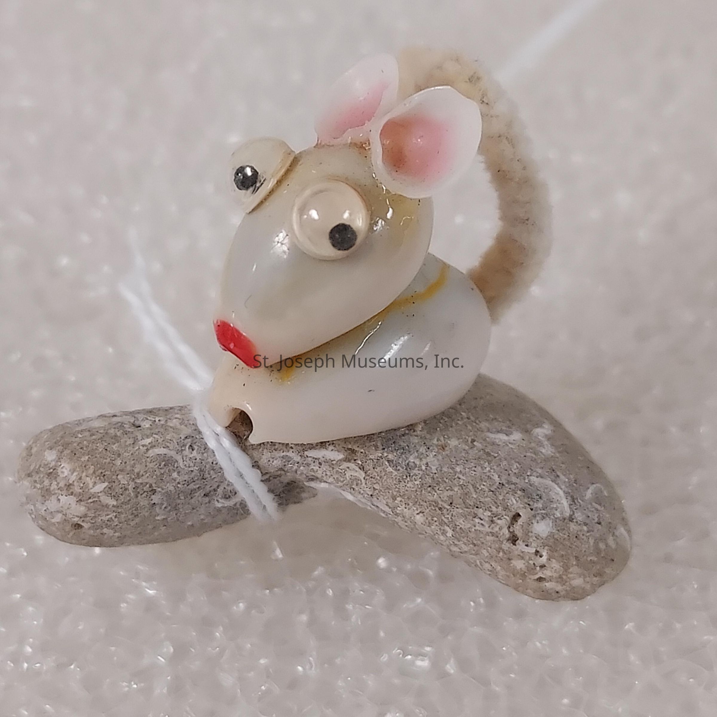 The image features a small, handcrafted mouse figurine made from seashells and stones, showcasing intricate and creative design. The mouse has a pair of conspicuous eyes, formed by smaller circular shells, and tiny pink ears, adding to its lifelike appearance. Its mouth is accentuated with a touch of red, giving a whimsical touch to the otherwise muted color palette. The mouse is perched on a pebble, which adds a base for the entire structure, and is tied in place with white thread that wraps around the stone. The background appears to be composed of a textured, white surface, enhancing the contrast and focus on the delicate figurine. The overall craftsmanship suggests careful attention to detail, emphasizing an artisan's skill in transforming natural materials into charming decorative pieces. This piece could serve as an intriguing, conversation-starter ornament for interior decor or as a unique artistic gift.