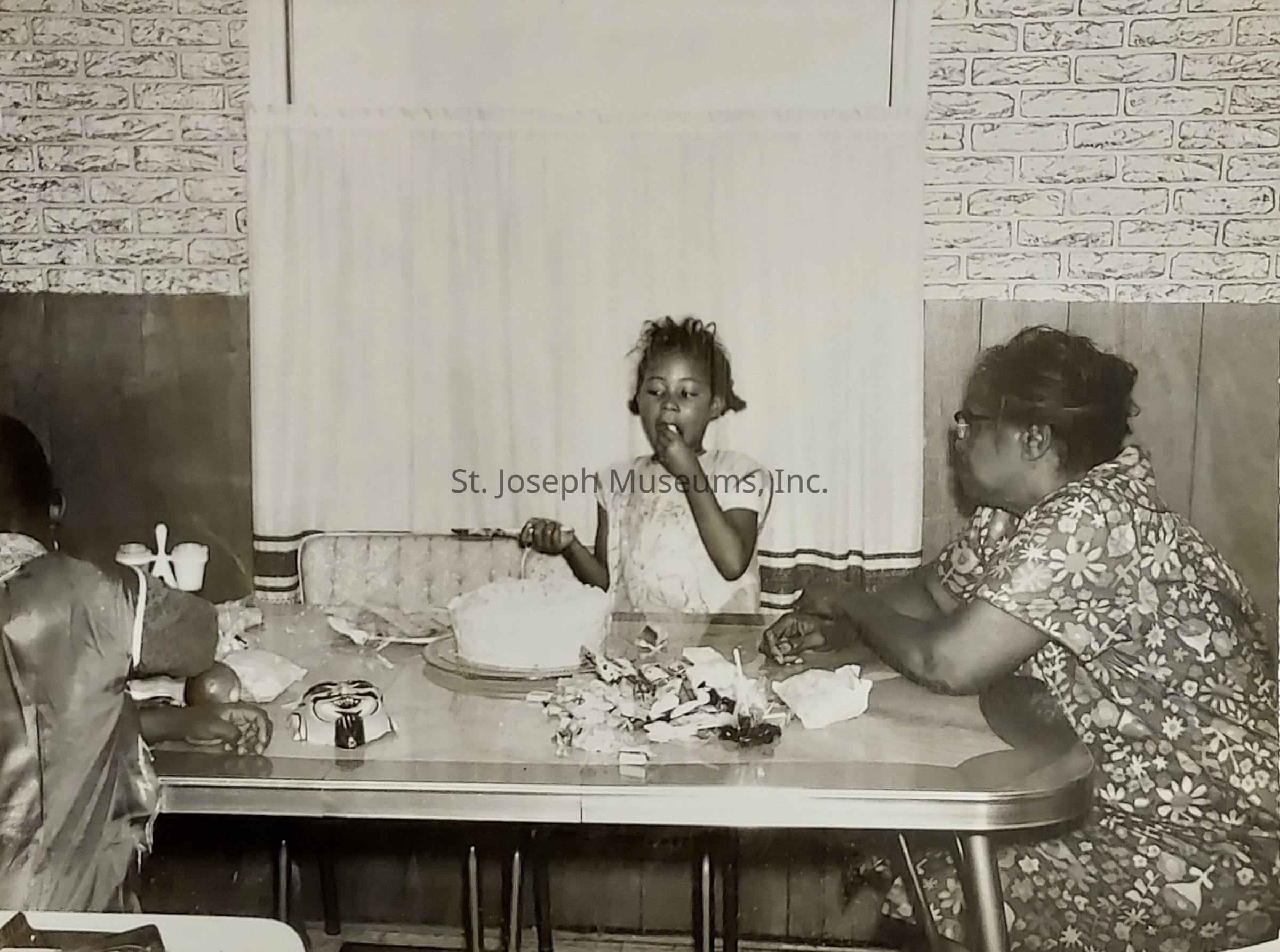 In this black and white photograph, a young girl is eating cake while standing at a table, surrounded by various small items and candy wrappers. To her left, someone with only their back visible is also seated at the table. On the right side, an older woman wearing glasses and a patterned dress is seated, leaning forward slightly, observing the girl. The setting suggests a warm, homely environment with patterned walls and a window adorned with curtains behind them. The photograph captures a candid, joyful moment shared among the individuals, highlighting a slice of everyday life.