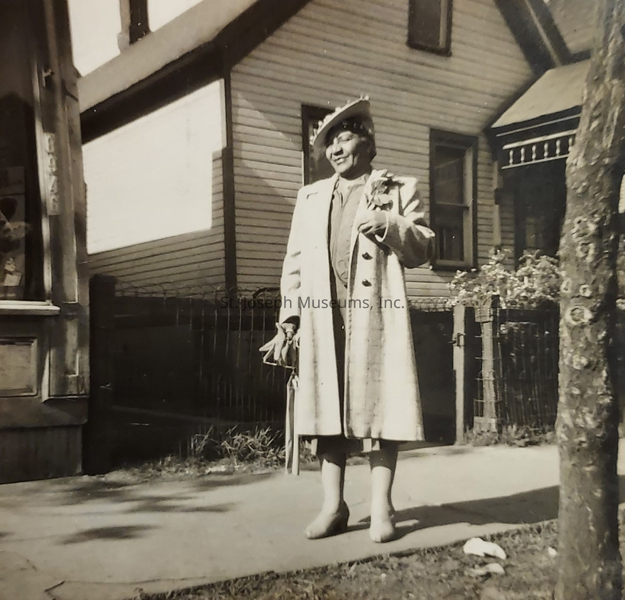 The black-and-white photograph features a well-dressed woman standing on a sidewalk in front of a house. She is wearing a long coat and a stylish hat, holding a purse and an umbrella in one hand. The house in the background is a two-story building with traditional architecture, characterized by its wooden siding and several windows. A small, ornate fence runs along the perimeter of the property, adorned with some greenery, suggesting a garden or yard area. The woman appears to be poised and elegant, suggesting that the photograph might have been taken during a special occasion or outing. Shadows cast by the tree and surrounding structures indicate the bright sunlight of daytime, contributing to the warm and nostalgic feel of the scene. Overall, the image captures a moment of timeless style and grace from an earlier era, offering a glimpse into the fashion and residential architecture of the time.