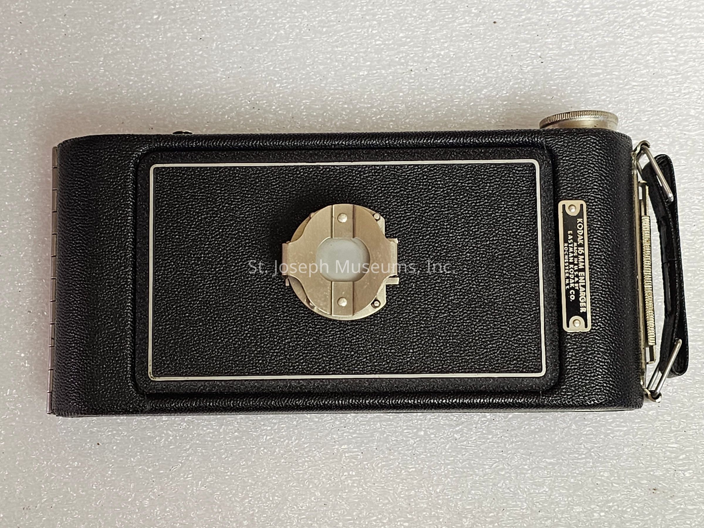 The image showcases a vintage camera, likely from the early to mid-20th century, with a compact and rectangular design, featuring a textured black exterior that provides a classic and durable appearance. The camera is lying flat against a white background, highlighting its sleek form and intricate details. At the center, there's a circular metallic component that might be part of the camera’s operational mechanism or a decorative element. A metal label on the right side displays text indicating the brand or model of the camera. Adjacent to this is a metallic knob, which probably serves as a control or a structural element. The design elements emphasize a blend of functionality and aesthetic appeal, characteristic of photographic equipment from that era, reflecting the technological and artistic sensibilities involved in the manufacturing of such items at the time. The entire setup serves as a nostalgic reminder of the evolution of photography and the craftsmanship involved in the production of cameras during the period when they transitioned from being luxury items to more accessible tools for documenting everyday life.