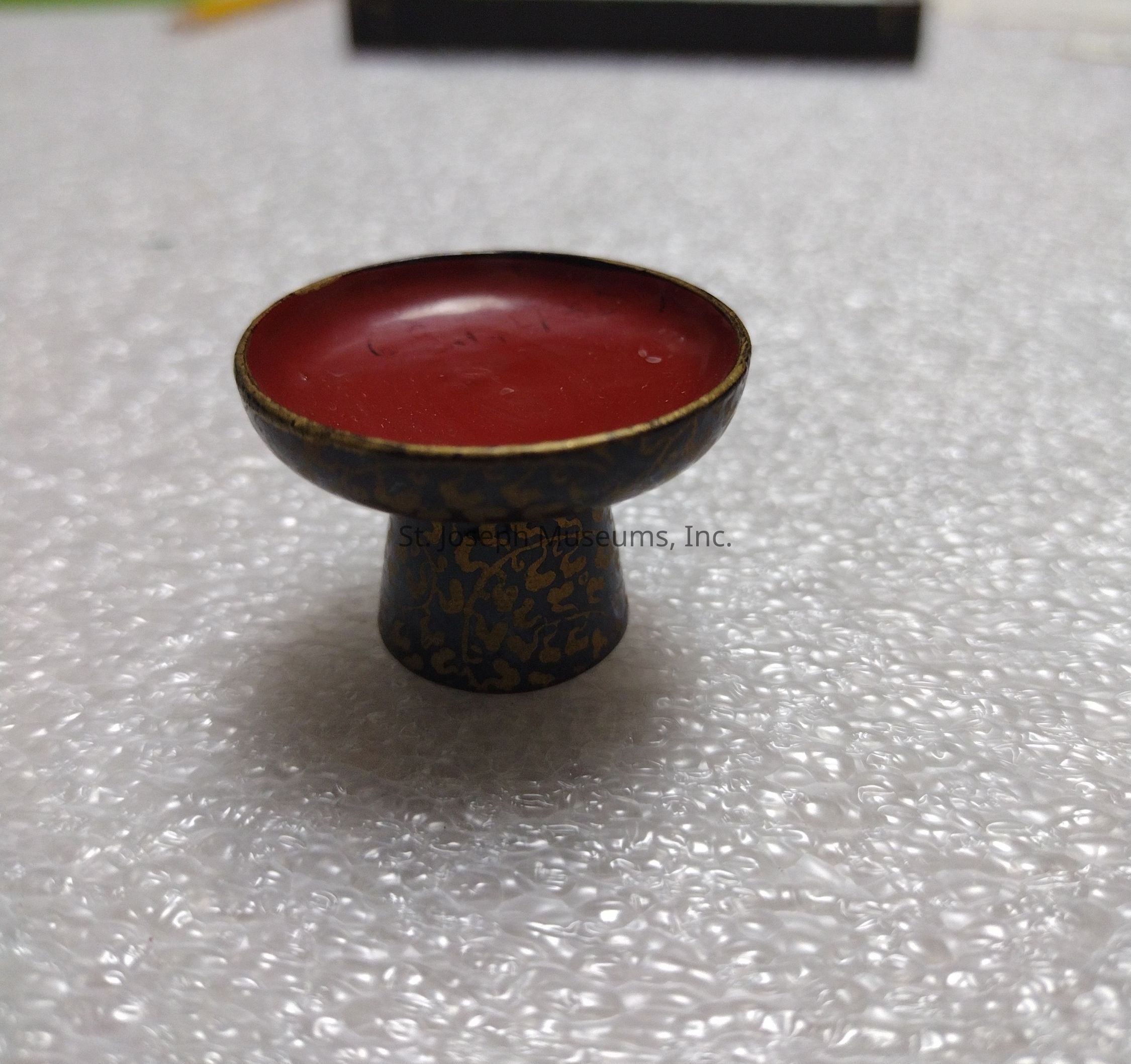 The image features a small, intricately designed chalice or goblet placed on a textured, bubble wrap-like surface. The chalice has a wide, shallow bowl with a rich red interior that contrasts vividly with its dark, detailed exterior, adorned with golden decorative patterns, possibly resembling vines or floral motifs. The base is a short, round stem that supports the bowl, creating a balanced and elegant appearance. The lighting highlights the glossy finish of the red surface inside the bowl, adding a touch of elegance and refinement. The textured background emphasizes the chalice's smooth curves and intricate design, making it appear as a significant artifact or decorative piece potentially used for ceremonial or ornamental purposes. The overall aesthetic of the chalice suggests a traditional or possibly cultural significance, characterized by detailed craftsmanship and a harmonious blend of color and texture.