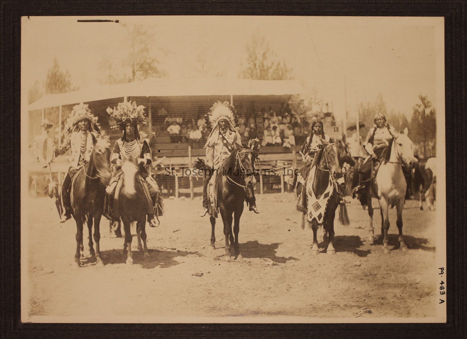 In the sepia-toned image, five individuals dressed in traditional Indigenous attire sit astride horses, each adorned with headpieces and regalia. They are positioned at the forefront of a scene that includes a background of wooden bleachers filled with spectators, suggesting an event is taking place. The headpieces, made of feathers and beads have intricate designs. The horses are embellished with decorative bridles and blankets. The setting is outdoor and rustic, with trees in the distance. The image portrays the individuals and their rich cultural expressions and also suggests community and identity being commemorated in a shared gathering.