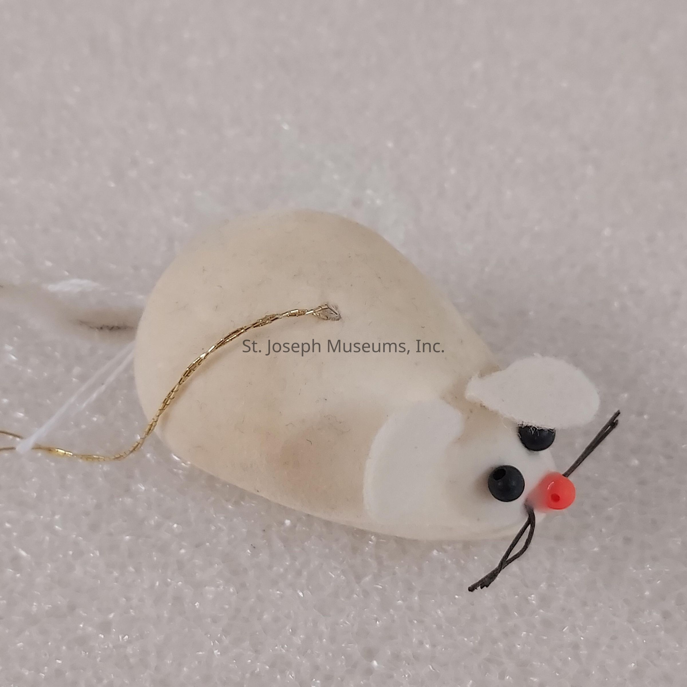 The image showcases a small, handmade mouse ornament with a white body, resting on a textured white surface. It features tiny black bead eyes and a red bead nose that stand out prominently against the light-colored fabric. The mouse is designed with delicate features, including round ears made of felt and slender whiskers crafted from thin black threads. A delicate, gold-colored string is attached to the mouse, suggesting it might be used as a hanging decoration. This ornament is carefully crafted to resemble a mouse, with an emphasis on simplicity and charm, making it a delightful decorative piece.
