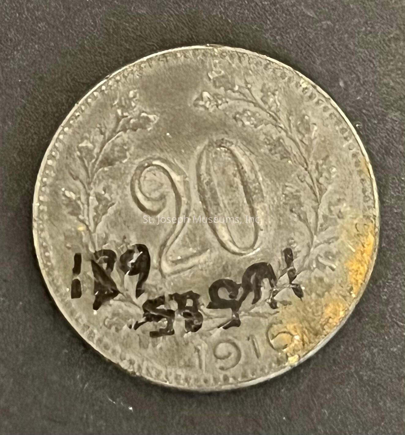 The image shows a vintage coin that appears to be weathered and has a denomination of 20 prominently displayed in the center. The coin has intricate detailing along the edges, including ornamental leaves or branches that curve to frame the numbers. Towards the bottom, there is a visible date that seems to read "1916." The coin's surface has patches of a yellowish discoloration, possibly due to age or exposure. The background is a dark surface that contrasts with the metallic nature of the coin, highlighting its tones and imperfections. This coin, despite its age and marks, carries a sense of history and craftsmanship, hinting at the period it originates from, while also presenting a unique juxtaposition of old and new with the addition of modern markings. The overall appearance suggests it might be a collectible item for coin enthusiasts.