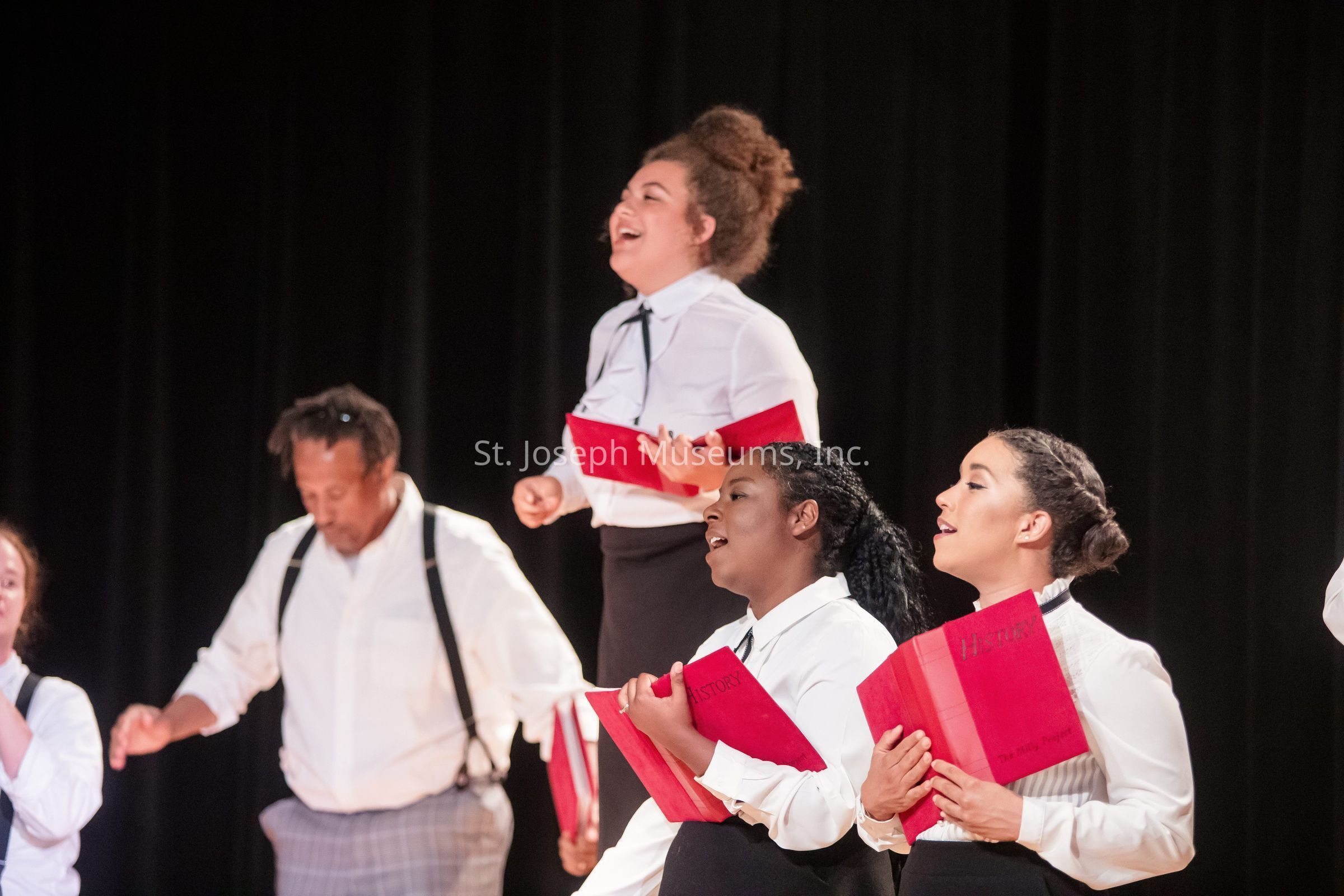 The image portrays a group of four individuals passionately singing during a choral performance. Dressed in formal white shirts, with some wearing suspenders and ties, they hold red hymn books, suggesting a traditional or formal setting, possibly for a choir performance. The expression on their faces reflects a deep emotional engagement with the music, signifying a moment of collective harmony and joy. Positioned against a dark background, the singers stand out vividly, with the lighting highlighting their enthusiasm and the intensity of the moment. The atmosphere seems to convey a sense of unity and shared purpose, with each person contributing their voice to create a harmonious sound. The presence of these books implies the performance involves historical or significant compositions. Overall, the image captures the essence of musical collaboration and the joy of shared artistic expression.