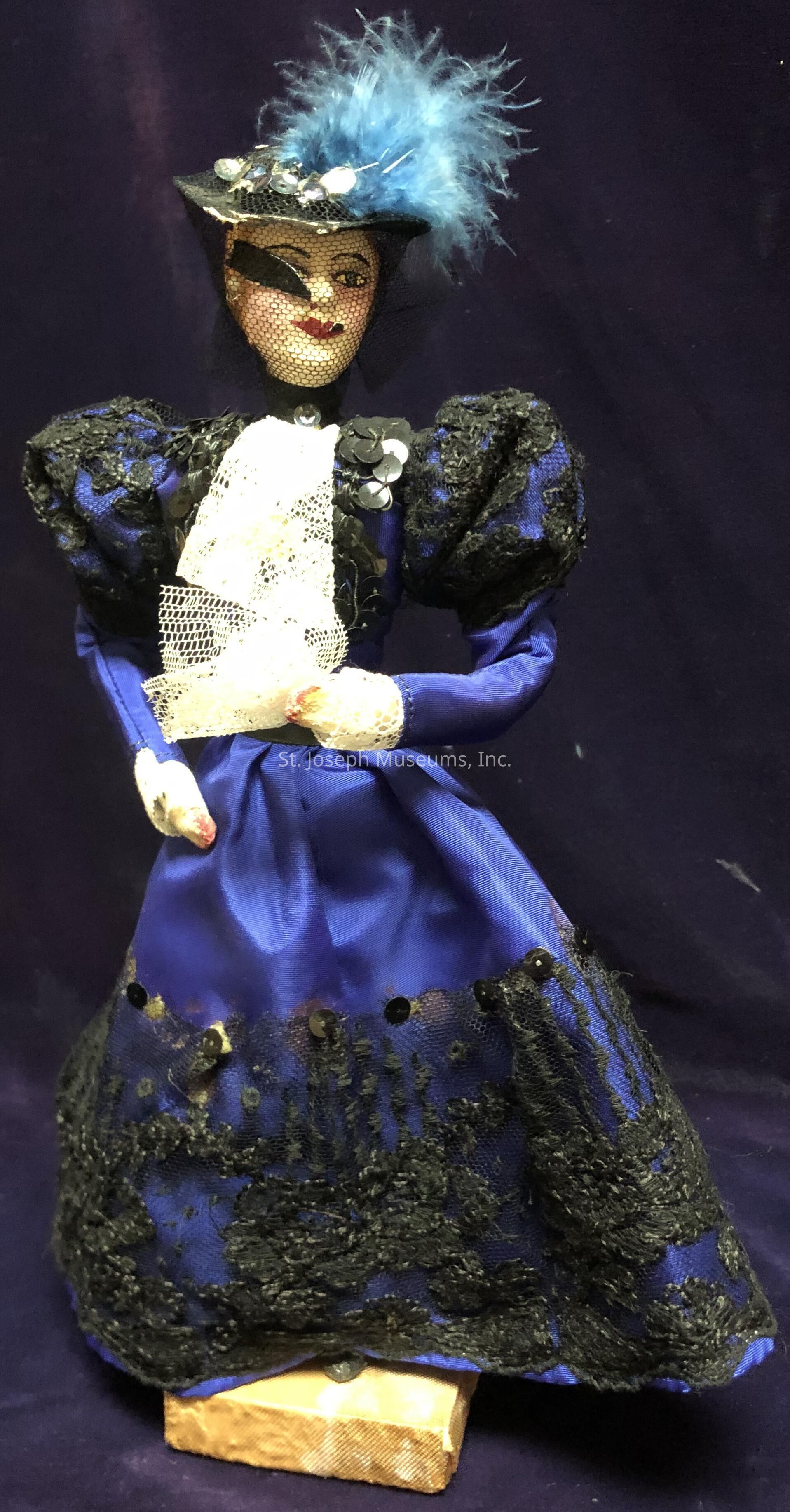 The image depicts a detailed doll dressed in elegant, vintage-style clothing. The doll wears a vibrant blue dress adorned with intricate black lace and sequins, giving it a luxurious and sophisticated appearance. A delicate lace shawl is draped elegantly over its front. The doll's face is painted with attention to detail, highlighting features such as red-toned lips and expressive eyes. It sports a stylish black hat, embellished with a striking blue feather, black netting, and shiny sequins. The doll stands on a small wooden block, set against a deep blue background that enhances the richness of its attire.