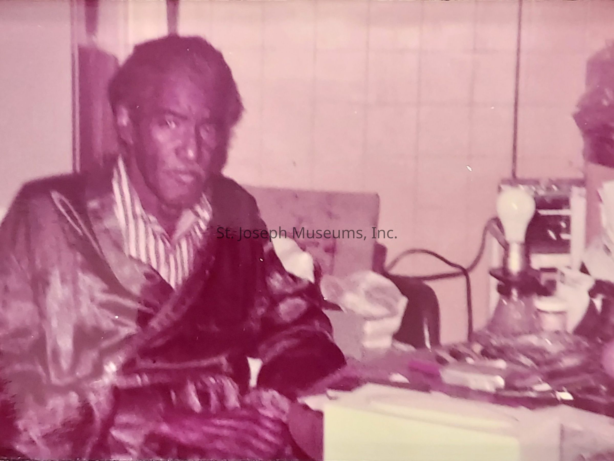 The image depicts a man sitting at a cluttered table in what appears to be an indoor setting. The man is wearing a striped shirt beneath a robe and is positioned in the foreground, looking toward the camera with a neutral expression. The table beside him is filled with various items, including a light bulb fixed in some sort of stand, scattered papers, and other miscellaneous objects, suggesting a workspace or a personal area. The background features tiled walls and some furniture, adding to the sense of an intimate, possibly lived-in space. The overall color tone of the photo gives it a somewhat vintage or aged appearance, adding character to the scene.
