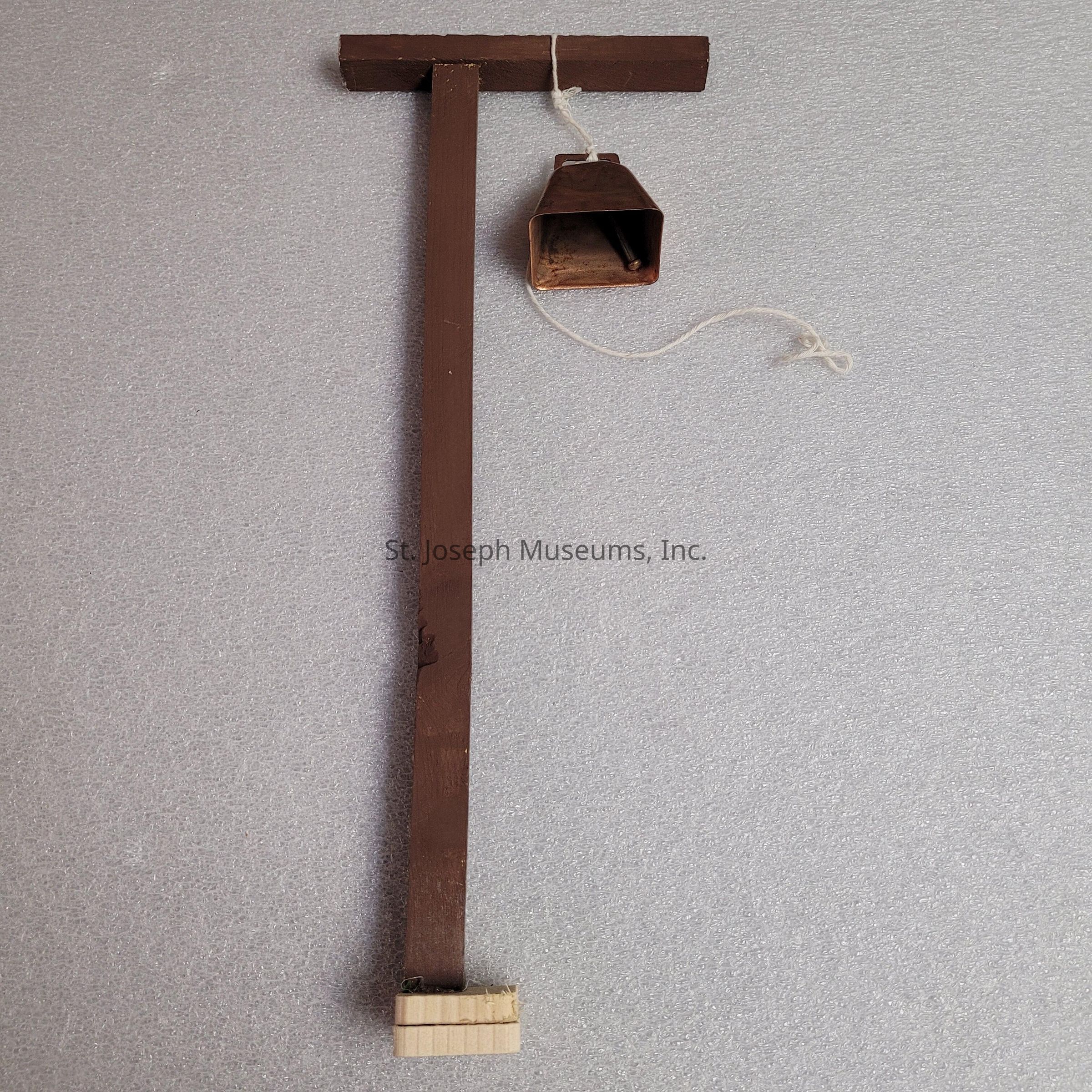The image depicts a wooden structure resembling a tall, narrow post with a T-shaped top, painted in a brown color. Attached to the top is a small, metal cowbell hanging by a piece of white string. The bell is slightly rusted, and the string dangles loosely from its top. The bottom of the wooden post has a small, rectangular block attached, appearing to provide stability or serve as a base. The entire setup rests against a light gray, textured background, giving an impression of simplicity and rustic charm. The overall composition suggests a handmade quality, possibly intended for decorative purposes or as a functional, minimalist design element.