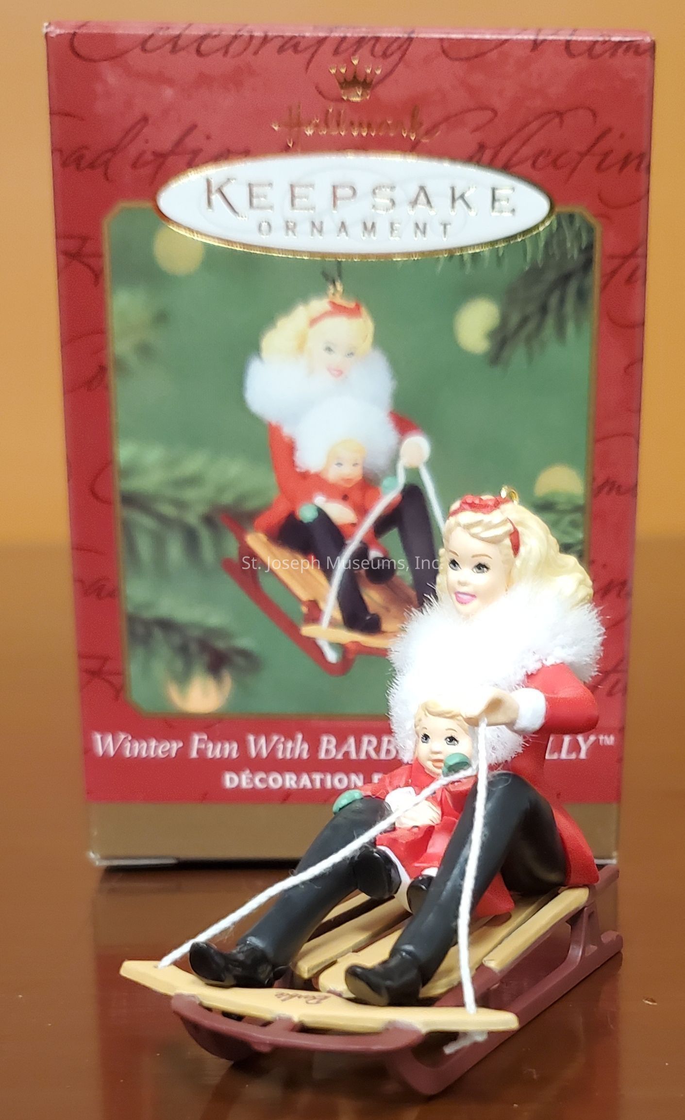 The image features a Hallmark Keepsake Ornament titled "Winter Fun with Barbie and Kelly," depicting a festive scene of a woman and a child on a sled. Both figures are dressed in red winter attire with white fur trim, suggesting a joyful winter activity. The woman is seated at the back of the sled, holding a rope, while the child sits in front, bundled warmly. This detailed ornament is vibrantly colored and placed in front of its packaging, which shows the same imagery, enhancing the winter theme. The box background is red, with gold accents and white text, giving it a holiday appeal.