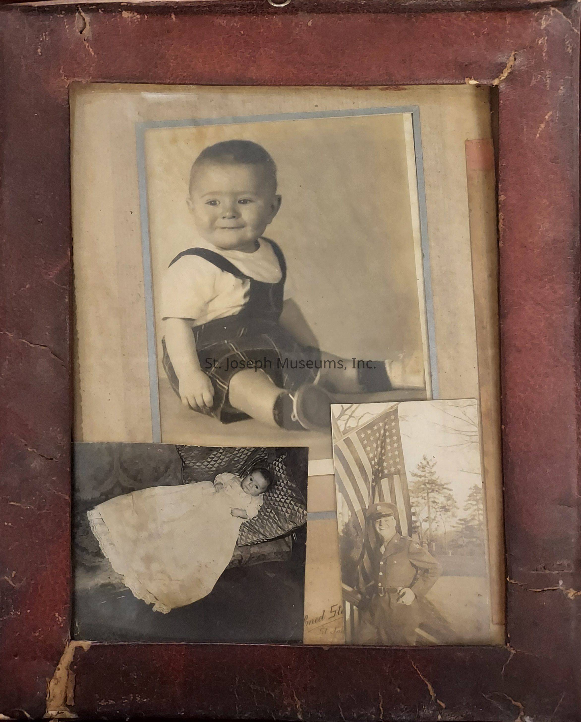The image shows an old, weathered photo frame holding three black and white photographs arranged closely together. The central picture prominently features a smiling baby dressed in a vintage outfit of a short-sleeved shirt with suspenders sitting comfortably. Below this, another photograph displays an infant lying on a patterned surface, wearing a long, elegant gown, presumably a christening or baptismal dress, which adds a classic touch to the image. On the right, the final photo depicts a person in uniform standing proudly with an American flag in the background, suggesting a scene of military significance or commemoration. The frame itself has a worn, reddish-brown appearance, showing signs of age with visible scuffs and some cracks, giving it a nostalgic and historical charm, perhaps indicating that these are cherished family memories or heirlooms spanning different significant life events and generations, each photo capturing distinct moments in time and hinting at the stories and heritage behind them.