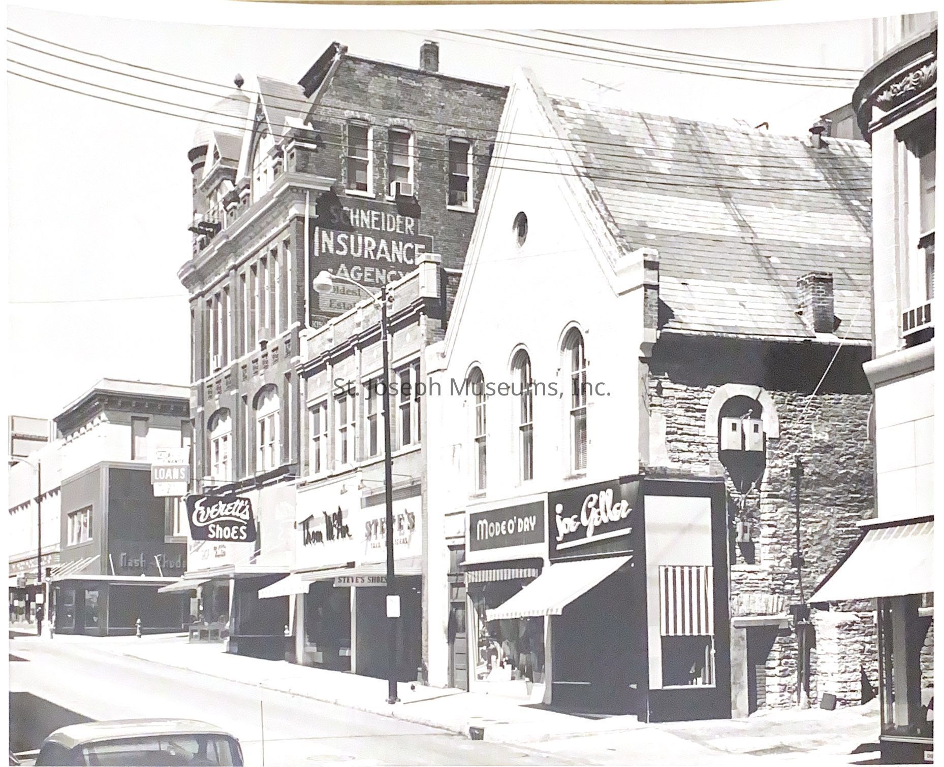 Black-and-white image depicting a row of storefronts, including Everett's Shoes, Thom McAnn, Steve's Shoes, Mode O'Day, and Joe Geller, situated on the north side of Felix between 7th and 8th. Above the buildings, there are signs for the Schneider Insurance Agency, and in the background, an old church with a steep roof and arched windows stands prominently.