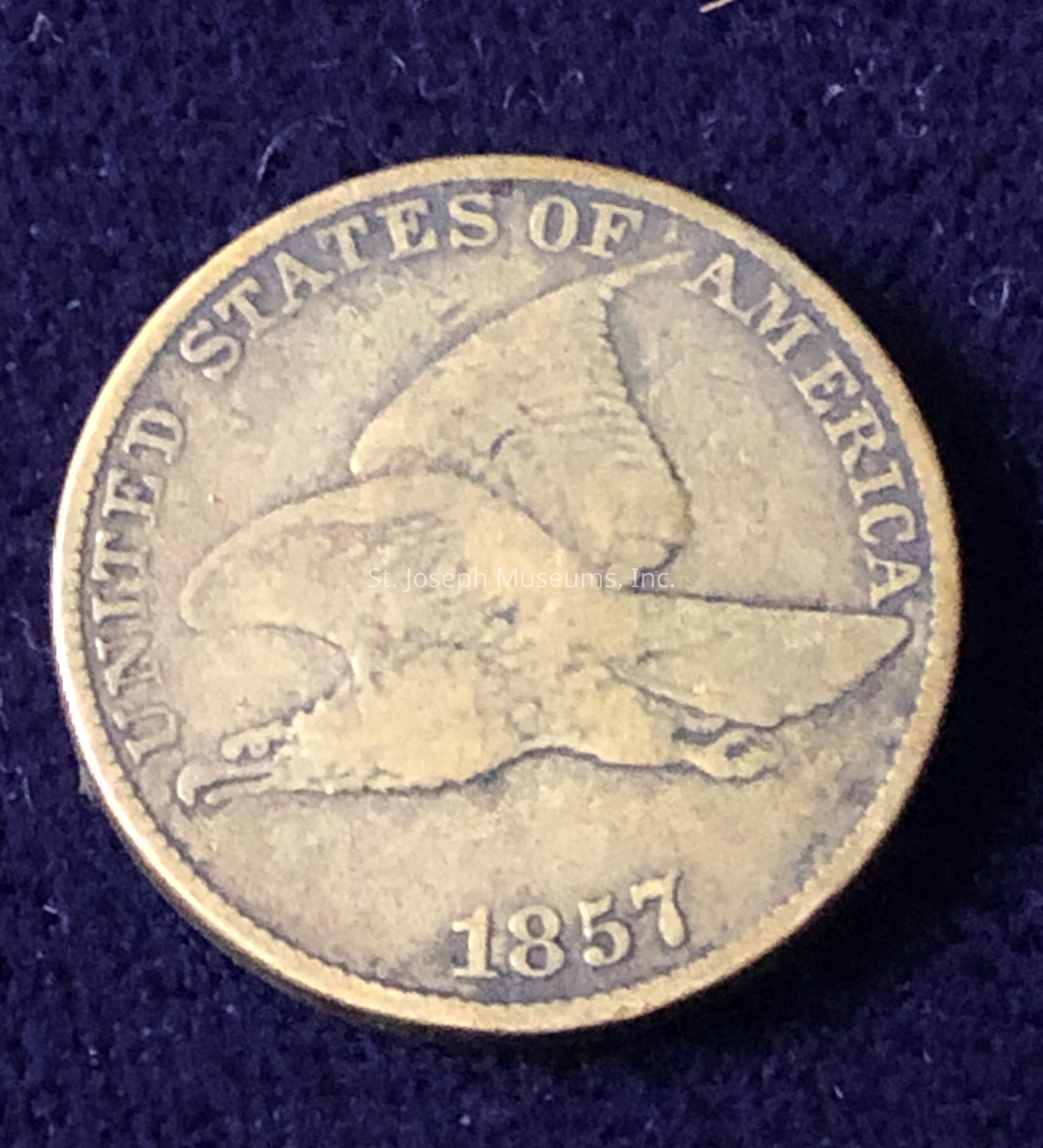 The image shows an old coin with a prominent design of an eagle in flight at its center. Around the edge is the inscription "UNITED STATES OF AMERICA," and the year "1857" is displayed at the bottom. The surface of the coin appears worn, indicating its age and frequent handling. The fine details of the eagle, such as its feathers and wings, are visible, providing a glimpse into the intricate craftsmanship of the era.