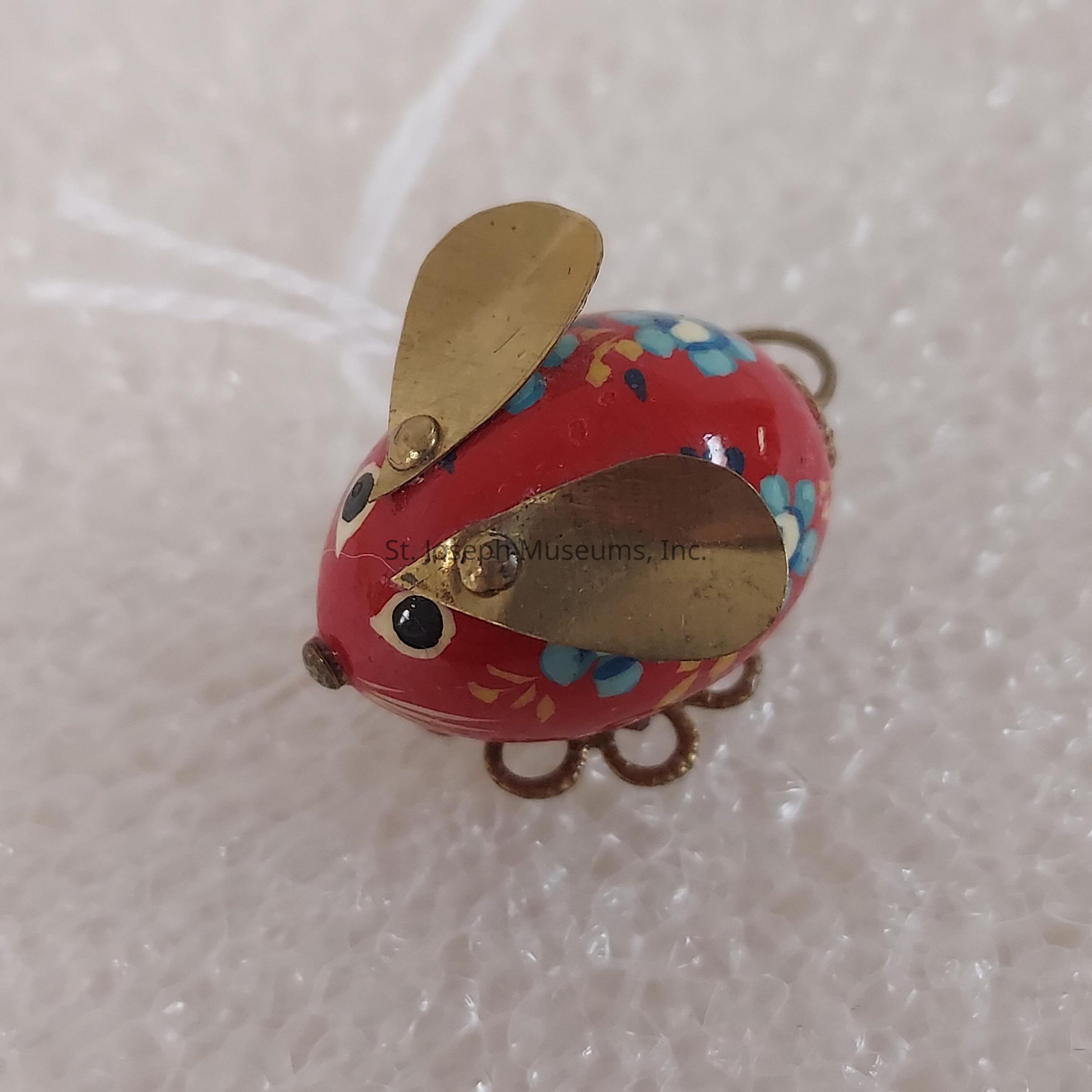 The image depicts a small, ornately crafted object resembling a mouse, featuring a charming red body adorned with floral patterns in blue and yellow, and a pair of large, rounded, gold-colored ears positioned on top. The mouse has black eyes that give it character, while its body appears to be made of a smooth, shiny material, possibly enamel. The base includes decorative elements that might represent feet or serve as a stand. The setting is a textured white background, likely a soft material, providing a neutral ground for the vibrant red and gold of the mouse to stand out, emphasizing its intricate design.