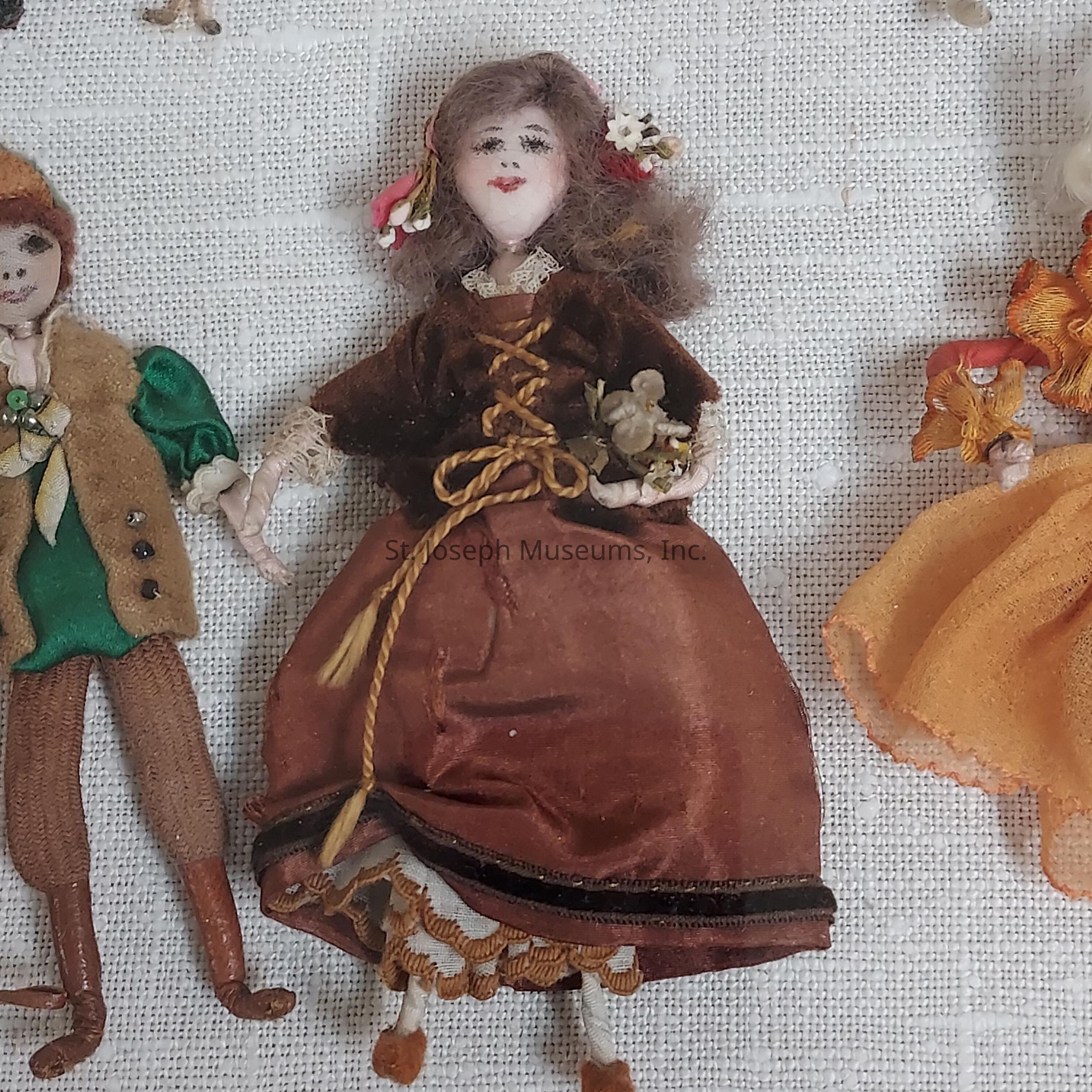 The image showcases three intricately designed dolls placed on a textured white surface. The central doll, dressed in a rich brown outfit with lace and floral embellishments, carries a small animal figure in her arms, and has long wavy brown hair adorned with flowers. To the left, another doll is seen wearing a green and brown ensemble, complete with a vest and hat, giving it a rustic appearance. On the right, a third doll is partially visible in a vivid orange dress, featuring lace and ribbon accents, suggesting a theme of vivid colors and detailed craftsmanship characteristic of vintage or handmade doll collections.