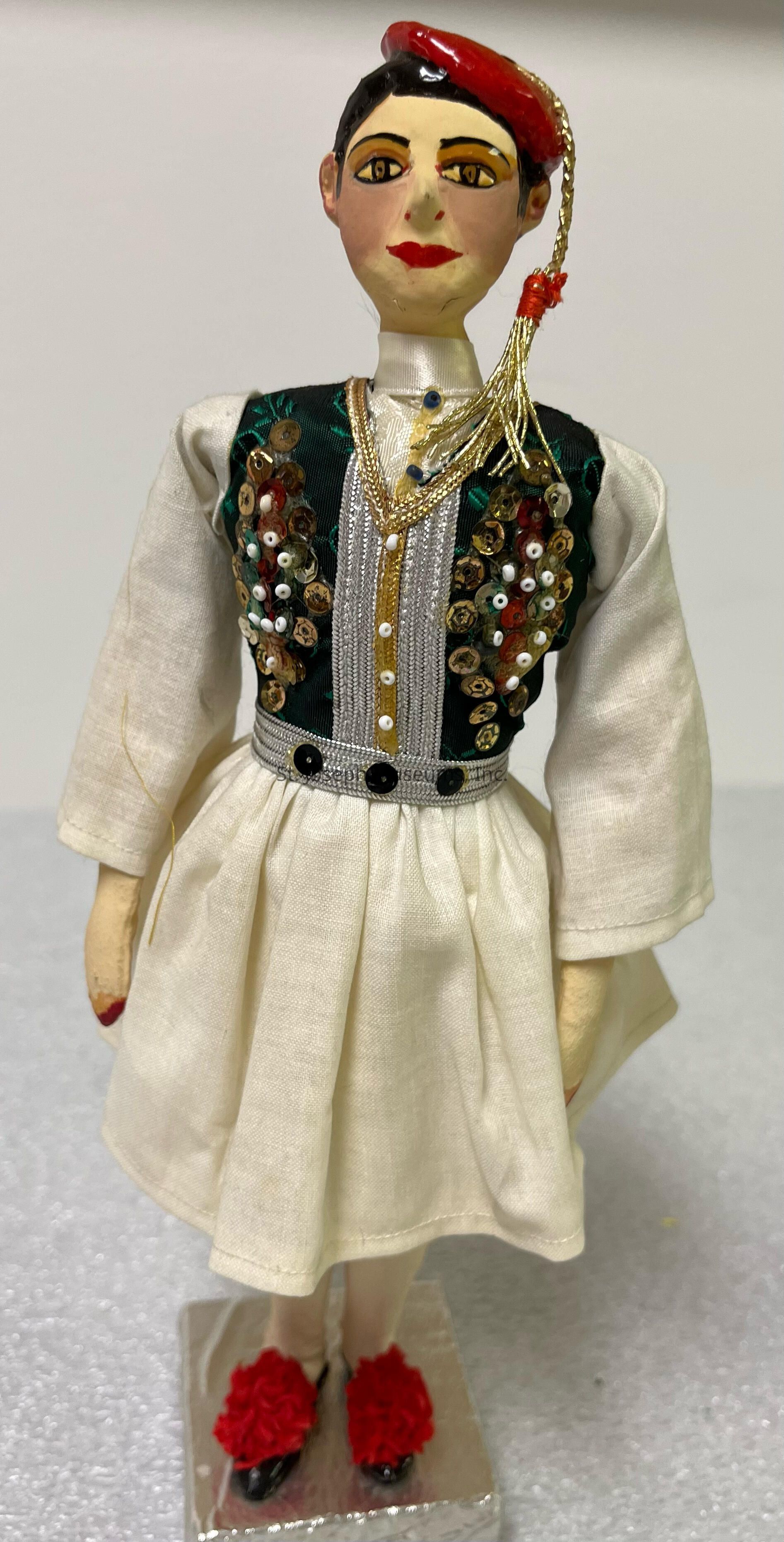 The image depicts a handcrafted doll dressed in traditional attire, featuring a white garment with wide sleeves and a pleated skirt. The doll's vest is elaborately decorated with sequins and embroidery in various colors, creating a pattern that feels rich and textured. A gold chain drapes over the front of the vest, adding a touch of elegance. The doll is topped with a red hat adorned with a gold and red tassel, adding to its cultural charm. Its shoes are black, embellished with vivid red pom-poms, providing an eye-catching contrast. The doll stands on a silver base, highlighting its decorative purpose.