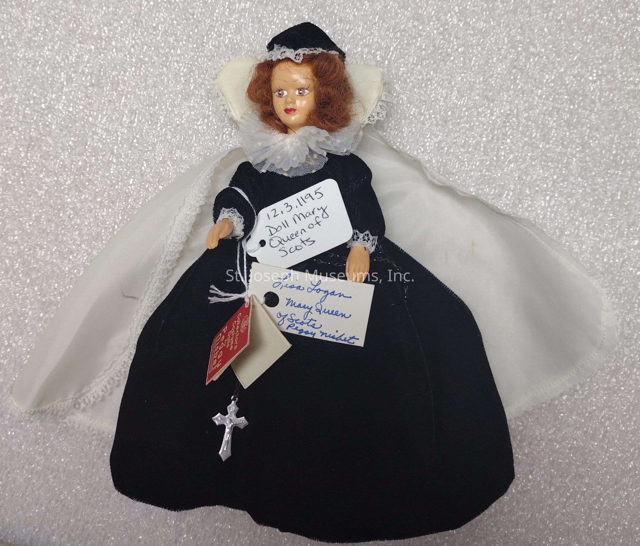 The image depicts a doll dressed to resemble Mary, Queen of Scots, with intricate historical attire in black fabric featuring a detailed lace collar and cuffs. The doll has reddish-brown curly hair and is adorned with a small silver cross pendant hanging from the outfit. Attached to the doll are several handwritten tags, with one noting the date "12-3-1195" and inscribed with "Doll Mary Queen of Scots." Another tag reads "Liza Logan, Mary Queen of Scots, Peggy Nisbet." The doll is laid on a textured white surface, which provides a contrasting backdrop, highlighting the elegant design and historical theme of the doll's attire and accessories.