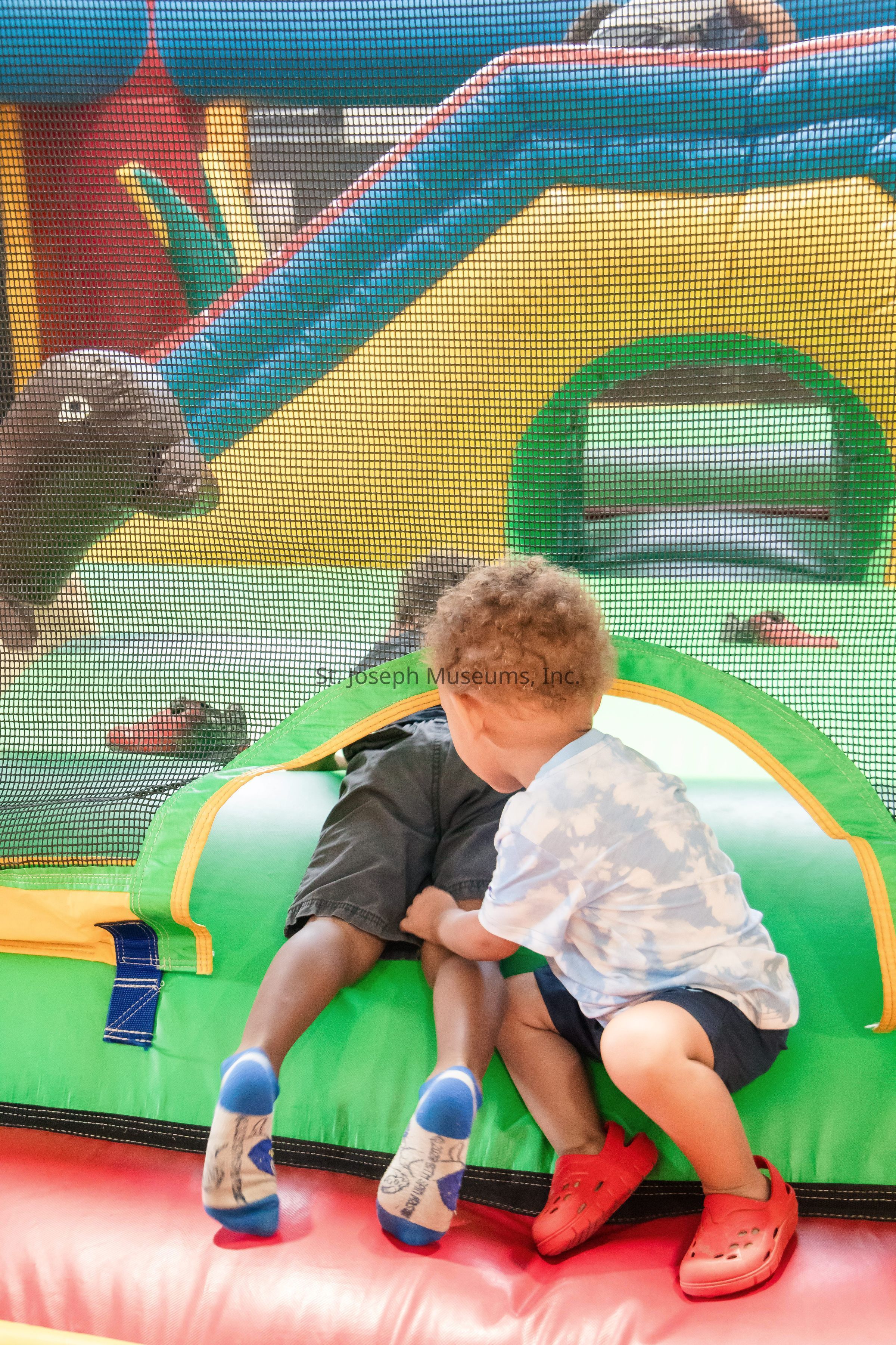 The image depicts two young children enjoying themselves in a colorful inflatable play area, with one child helping the other to either enter or exit a small circular opening; the vibrant setup features blue, green, yellow, and red elements, and a mesh safety net in the background ensures a secure environment for the playful activities, highlighting themes of friendship, cooperation, and innocent joy in a lively, engaging setting designed for children’s amusement and interaction.