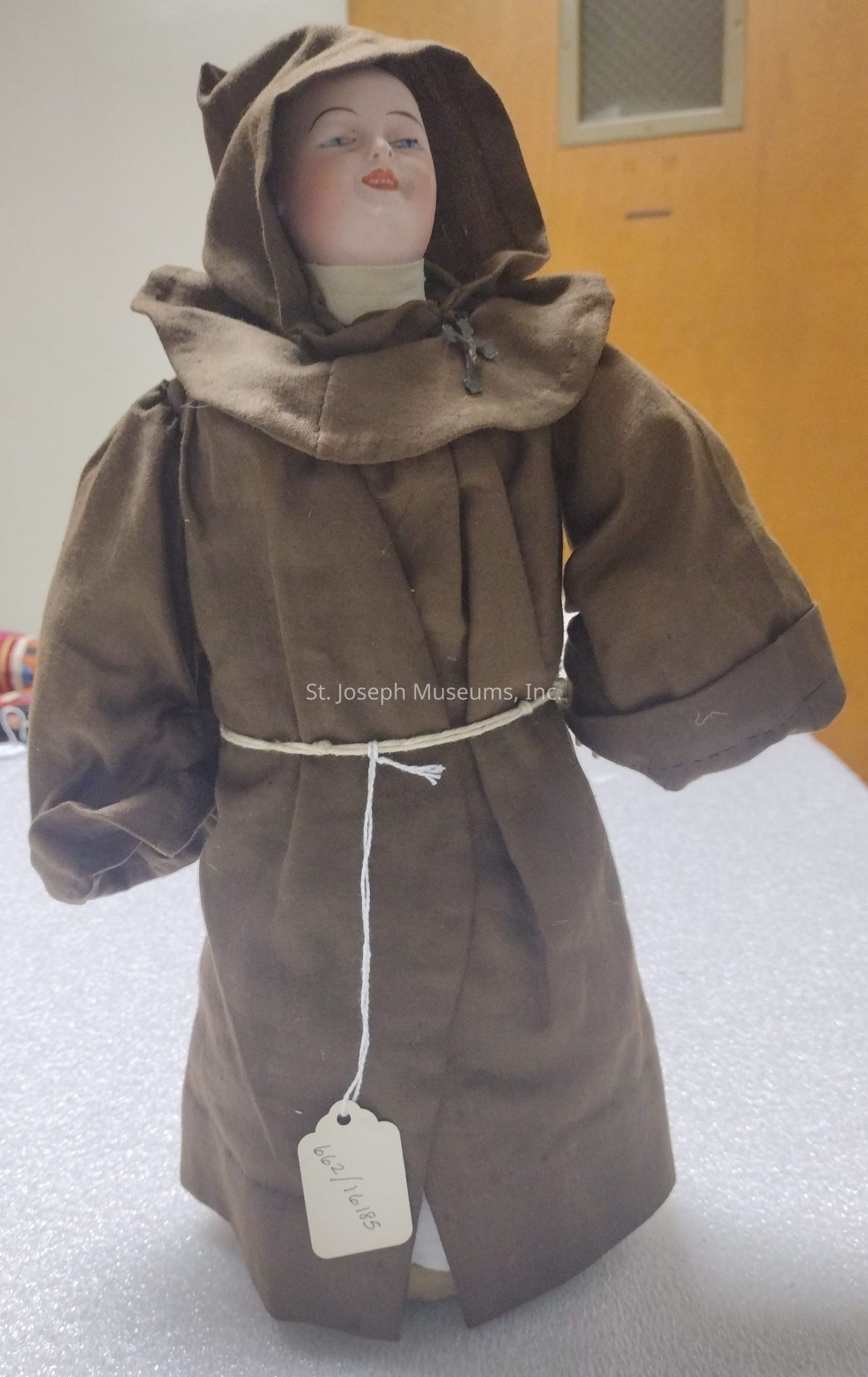 The image shows a doll dressed in a brown monk-like robe with a hood, featuring a tied string around the waist and a tag hanging from it. The doll's face is painted with delicate features, including a serene expression and rosy cheeks, while its hands are hidden within the robe's long sleeves. This setup gives the impression of a quaint and historical piece, possibly handmade or crafted to resemble a figure of monastic life. The background features a plain setting with a wooden element in the upper part, providing a simple and undistracting backdrop to the doll.