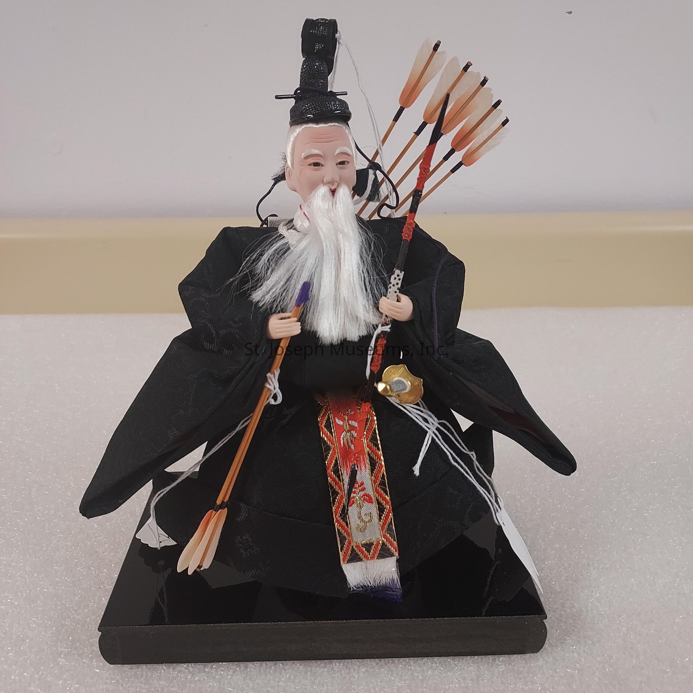 The image depicts a traditional Japanese doll intricately crafted, showcasing a figure with a long white beard and mustache, wearing an elaborate black robe adorned with intricate patterns and a colorful sash featuring red and gold designs. The figure has a serene expression, and atop its head, it wears a black headpiece. In its right hand, the doll holds several arrows with feathered tips, while in its left hand, it grasps a bow, indicating its archer identity. The doll is seated on a shiny black base, which complements its elegant attire, emphasizing the craftsmanship and cultural significance typical in such traditional Japanese dolls, often regarded as collector's items or symbolic artifacts representing historical and mythological figures.